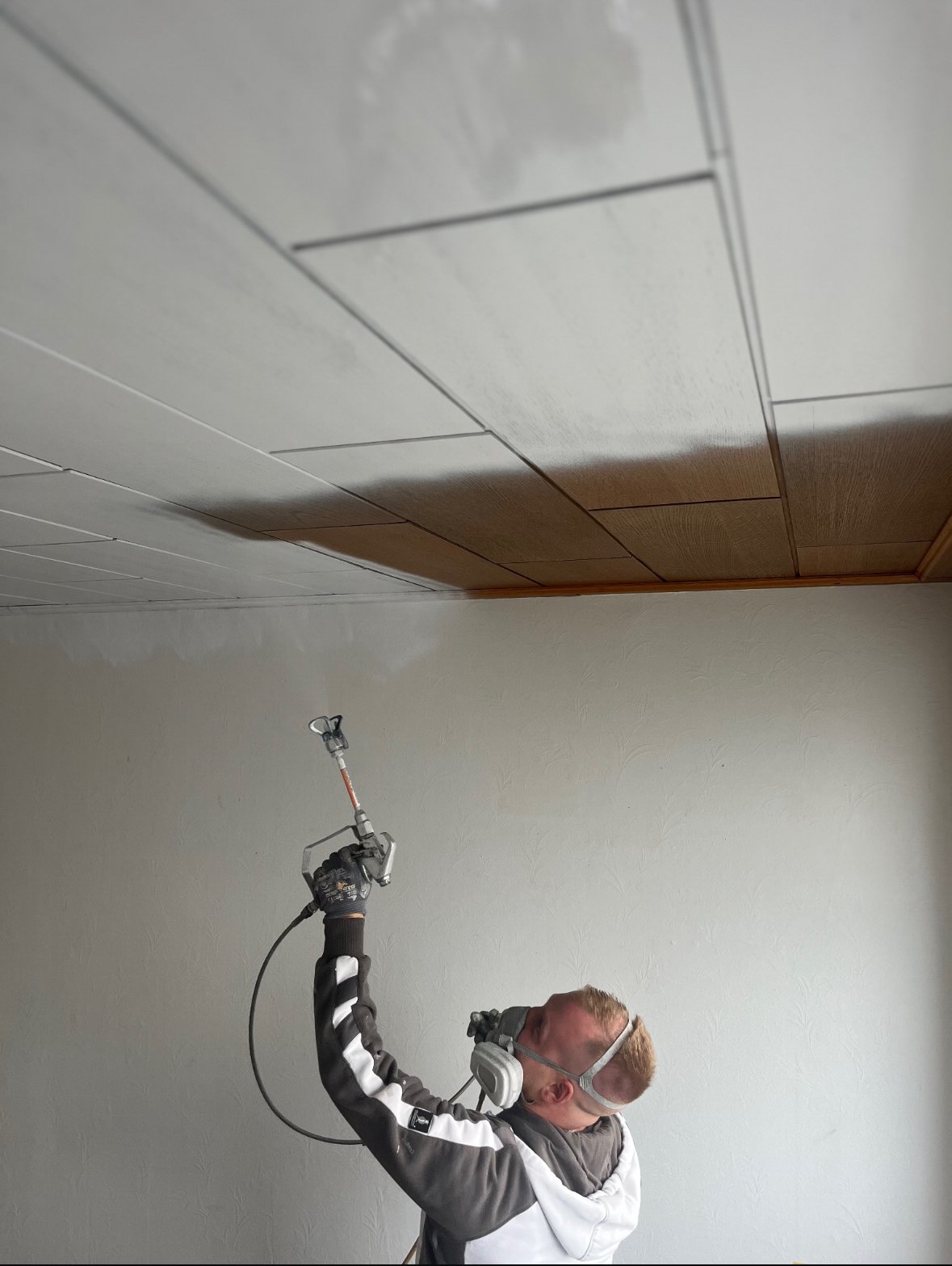 Mann bei der Arbeit mit einem elektrischen Werkzeug an der Decke eines Raums, trägt Schutzbrille und Gehörschutz, weiße Wand im Hintergrund.