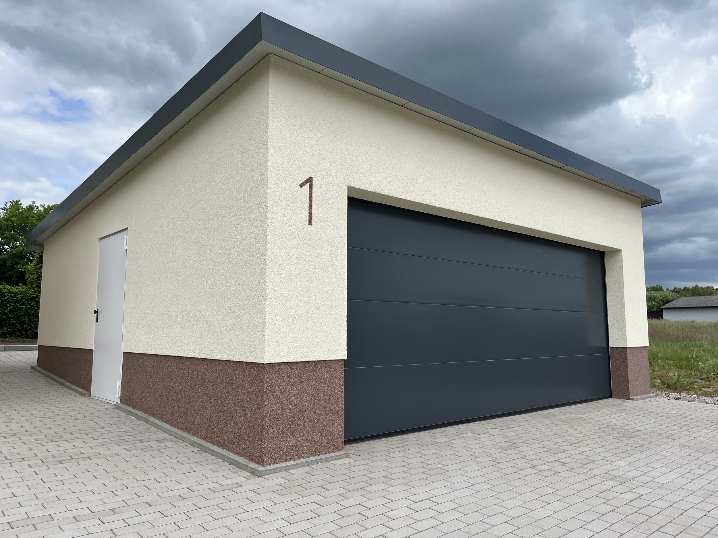 Ein moderner Carport mit dunkelblauer Tor- und Seitentür, nummeriert mit '1', auf gepflastertem Boden, vor Himmel mit dunklen Wolken.