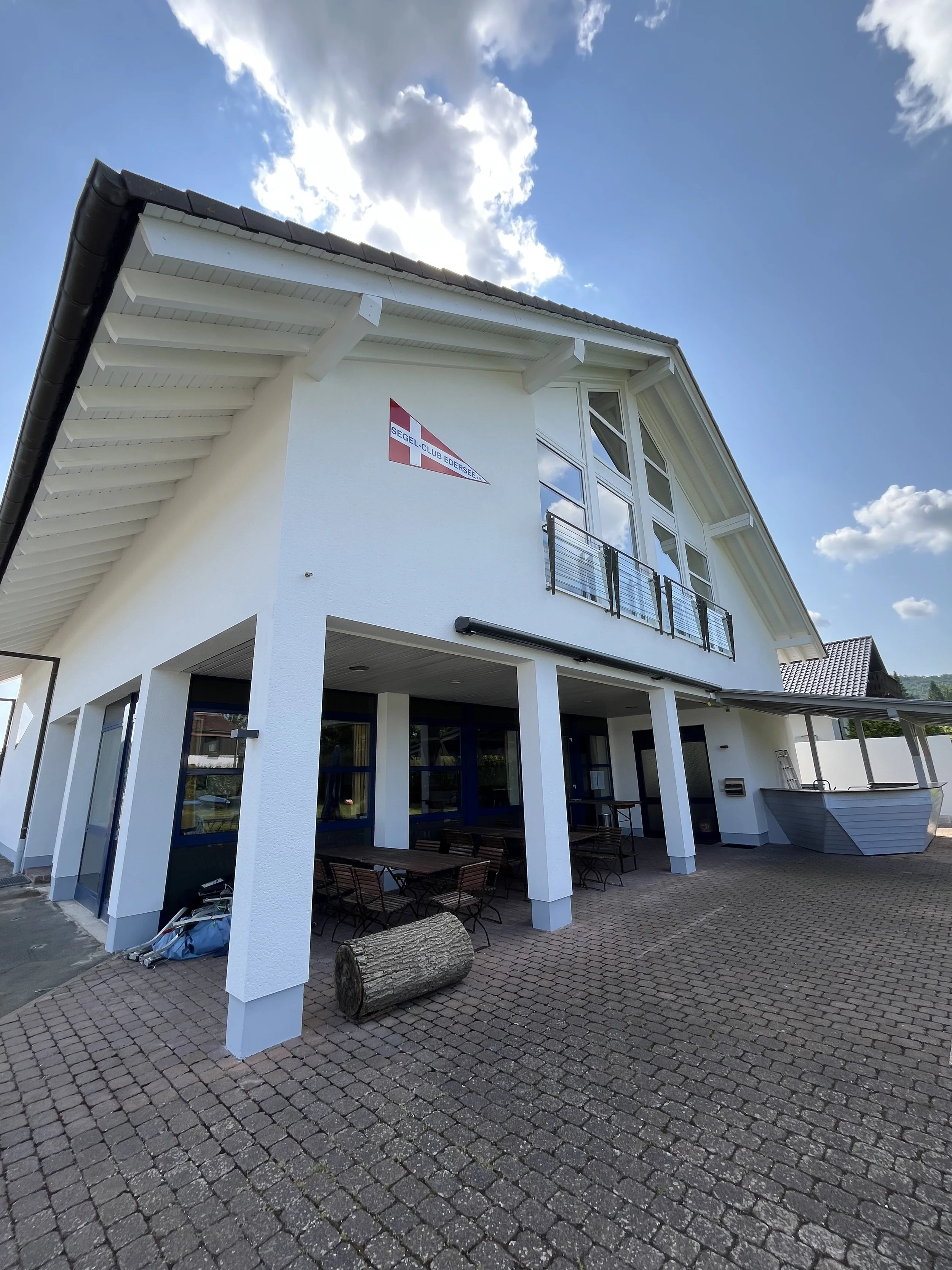 Weiße mehrstöckige Gebäude mit großen Fenstern, Balkon und einer Terrasse im Freien. Es gibt ein Logo des Segel-Club Edersee an der Wand. Der Himmel ist teilweise bewölkt, und der Boden besteht aus Pflastersteinen.