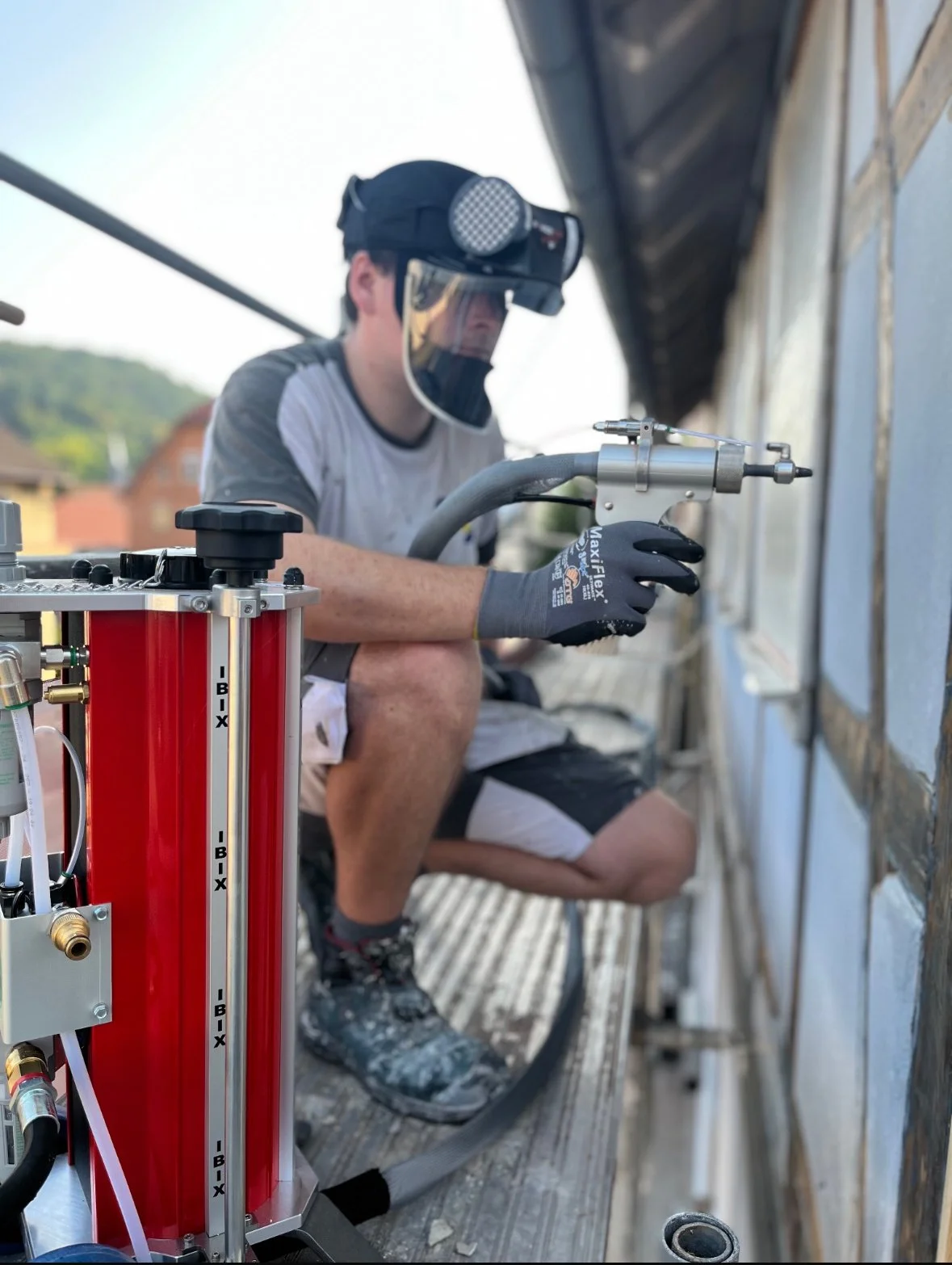 Mann trägt Schutzhelm und Handschuhe, arbeitet mit einem Heißluftbrenner vor einem Gebäude, vermutlich im Rahmen einer Haus- oder Fassadenrenovierung.