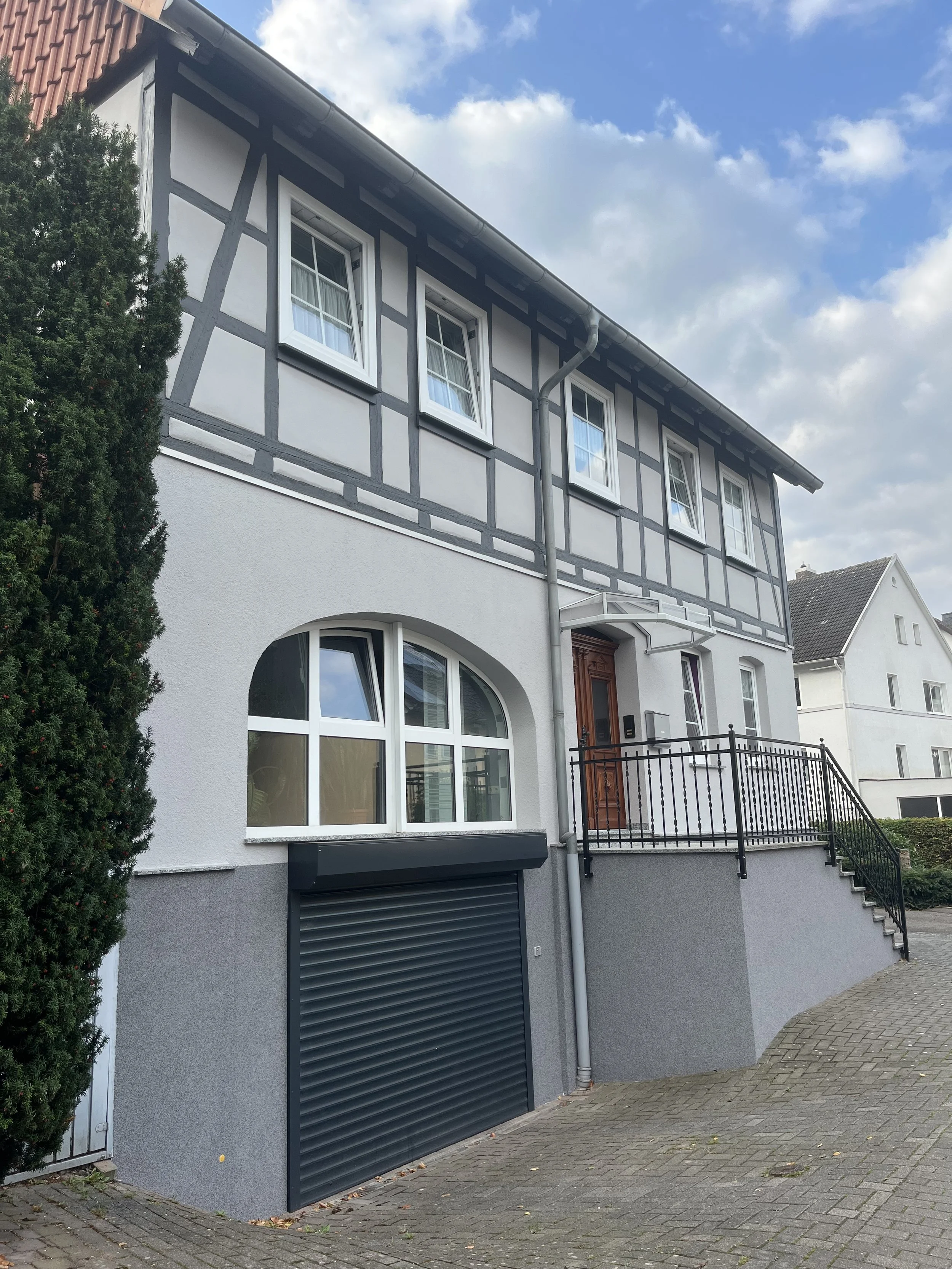 Mehrstöckiges Haus mit einer Garage im Erdgeschoss, einem Eingang mit Treppe und einem Balkon, im Fachwerktil, mit grauer und weißer Fassade, unter einem bewölkten Himmel.