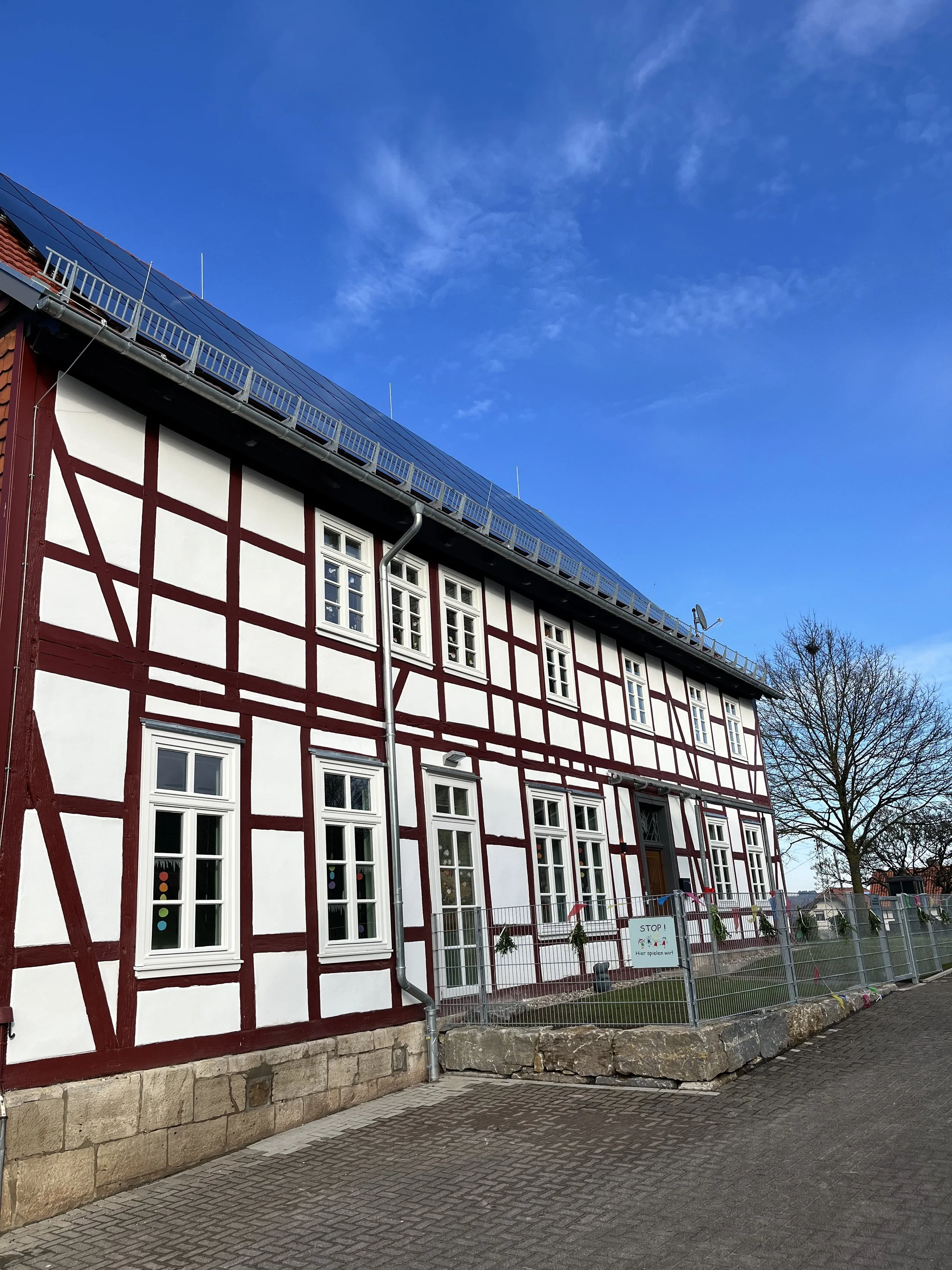 Ein Gebäude im Fachwerkstil mit weißen Wänden und roten Holzbalken, Dach mit Solarmodulen, Baum neben der Fassade, blauer Himmel mit wenigen Wolken.
