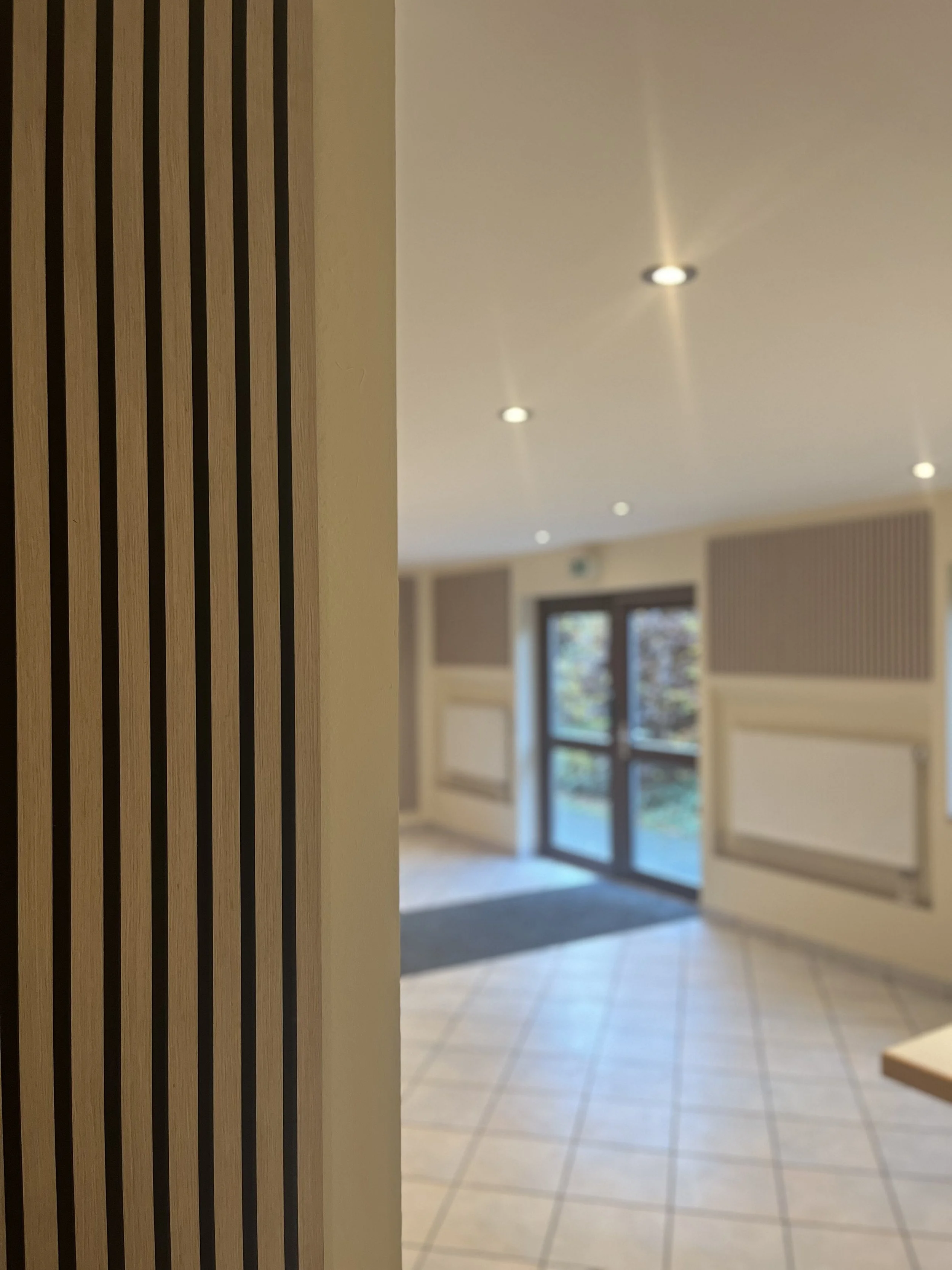 Innenaufnahme eines Flurs mit einer Holzwand mit vertikalen Lamellen, Fliesenboden, weißen Wänden mit Wandradiatoren, einem Fenster und einer Glastür, Deckenleuchten.