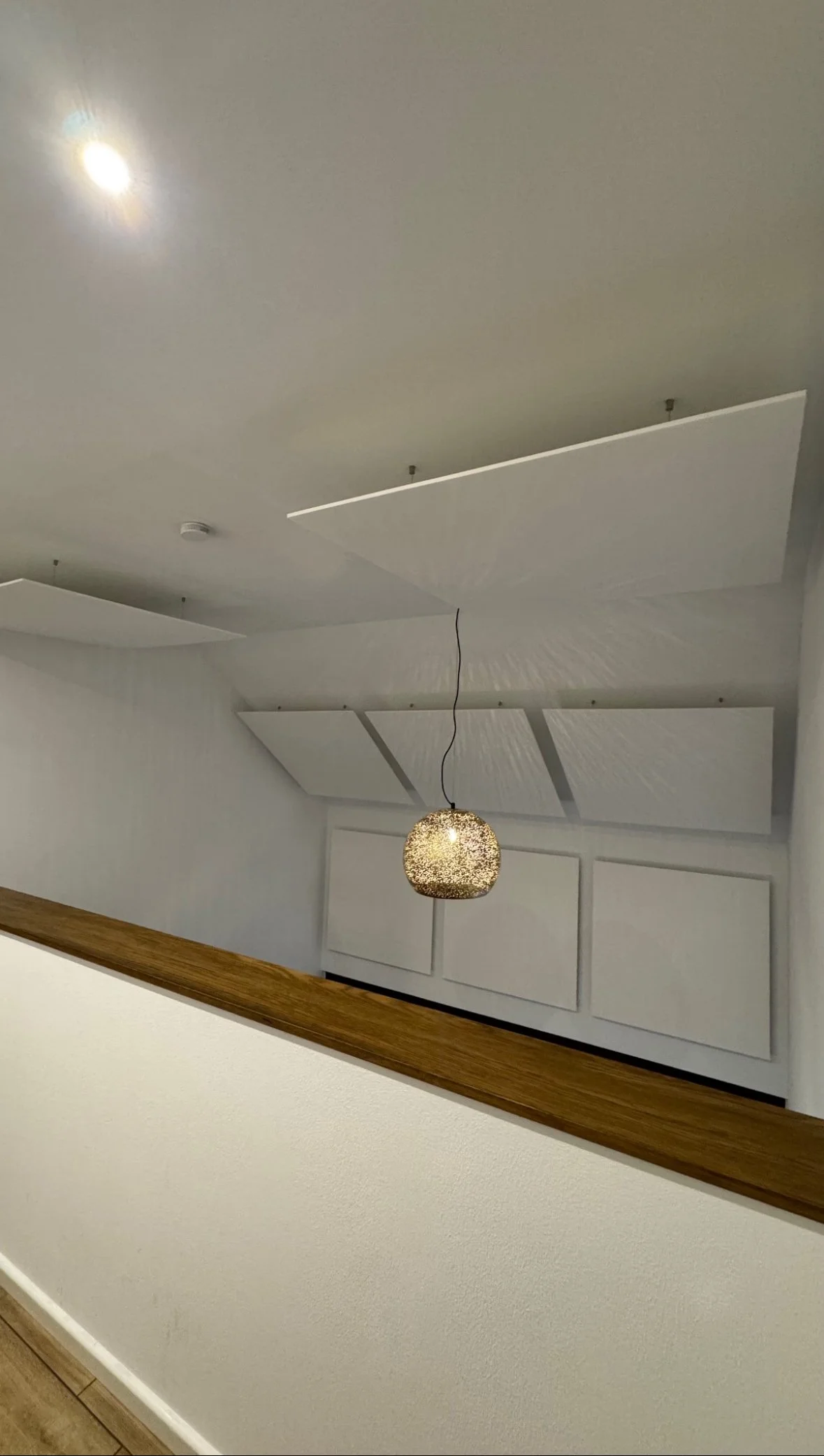 Innenansicht eines Gebäudes mit weißen Wänden, sechs weiße Deckenpaneele, einem bedeckten goldfarbenen Hängeleuchter und einer Holzgeländer