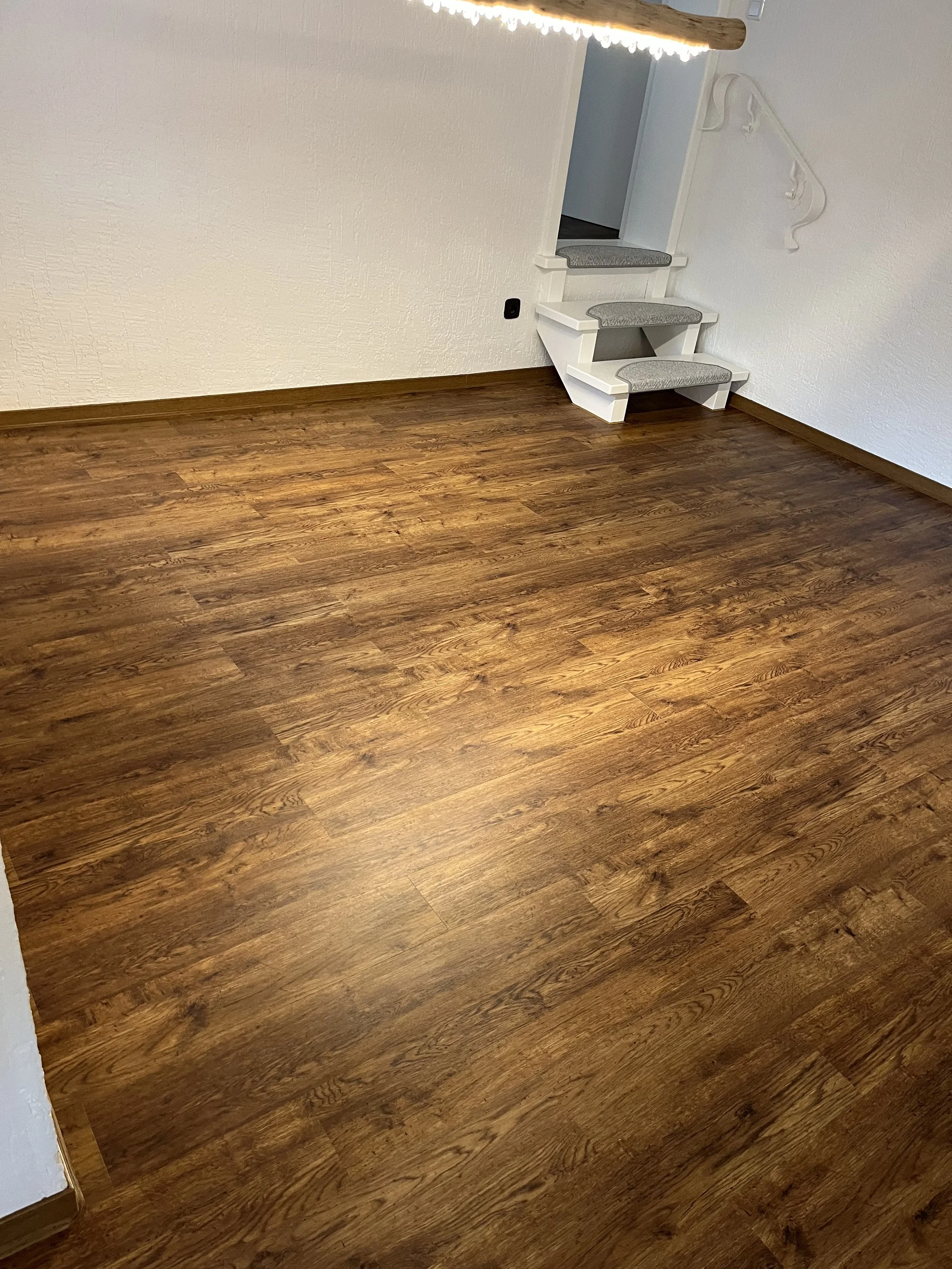 Ein Raum mit holzfarbenem Laminatboden, weiße Wände und eine kleine Treppe mit grauen Teppichstufen.