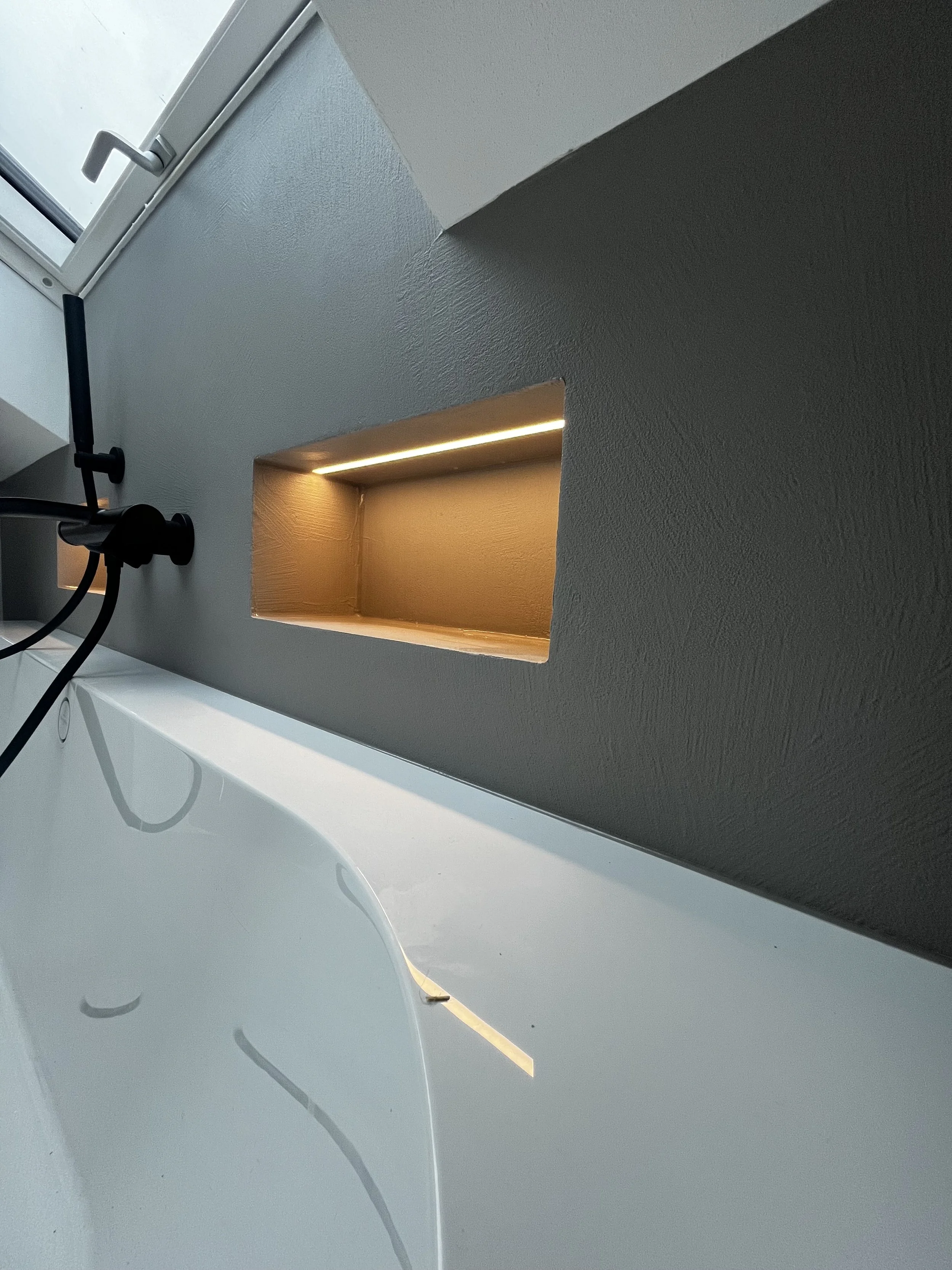 Ein weißes Bad mit einer Badewanne, an der Wand ist eine rechteckige Nische mit Beleuchtung. Ein schwarzer Duschkopf hängt an der Wand.