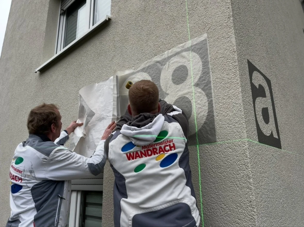 Zwei Männer in weiß-grau Arbeitsjacken bei der Anbringung eines grafischen Films auf einer Hauswand, neben einem Fenster. Es scheint eine Wandkunst oder ein Beschriftung zu sein.