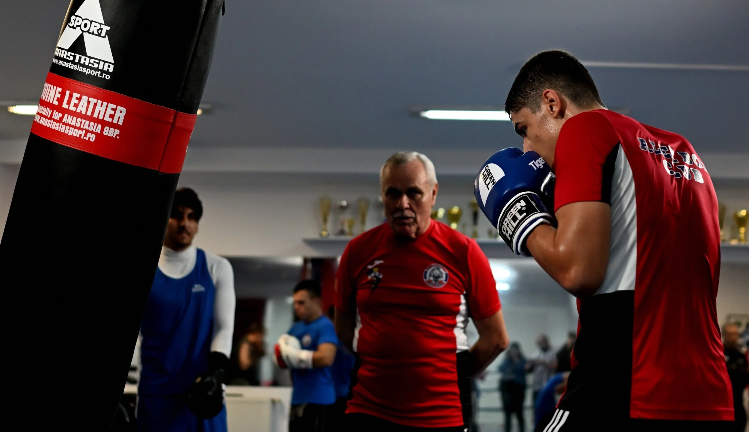 A boxing gym with boxers and a coach, one boxer in red gloves and shirt, another in blue gear, and a coach in a red shirt, with a punching bag on the left.