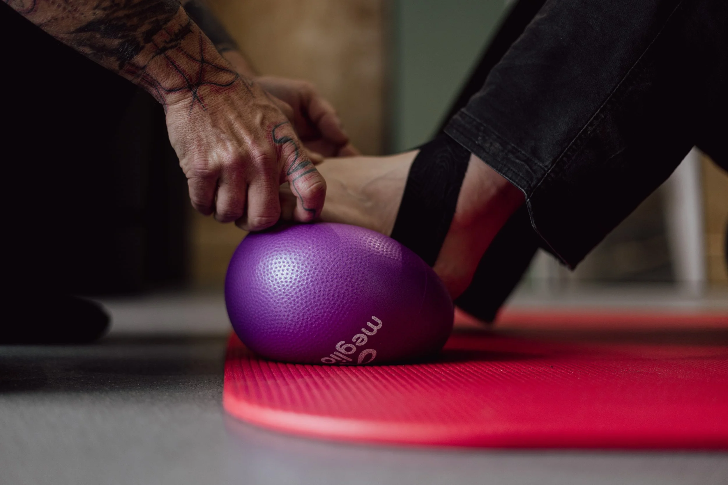 A person wearing black trousers is rolling out a foot with a purple exercise ball on a red yoga mat, being assisted by a physio therapist.