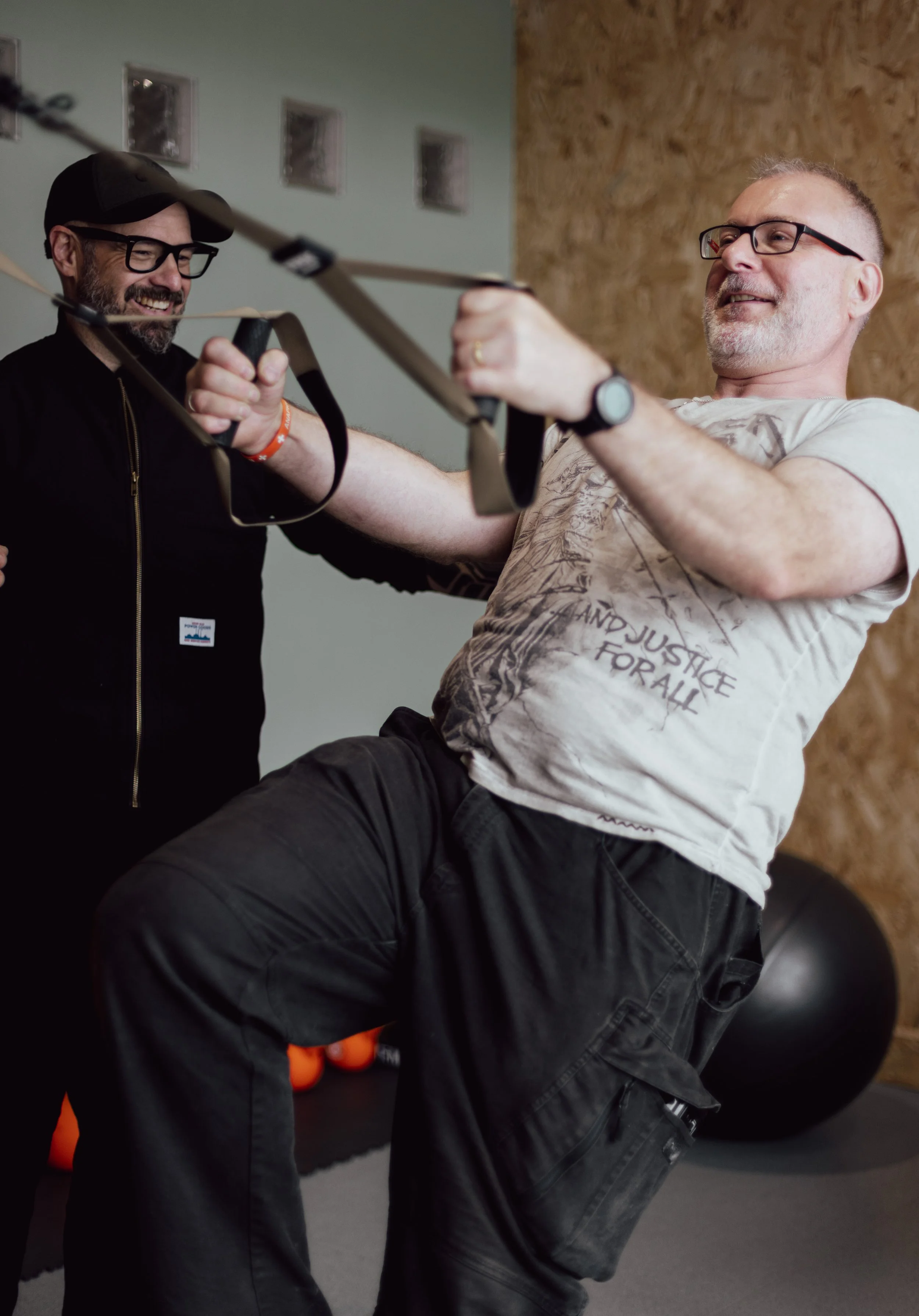 Two men in a gym engaging in a training activity with resistance bands, smiling and laughing.
