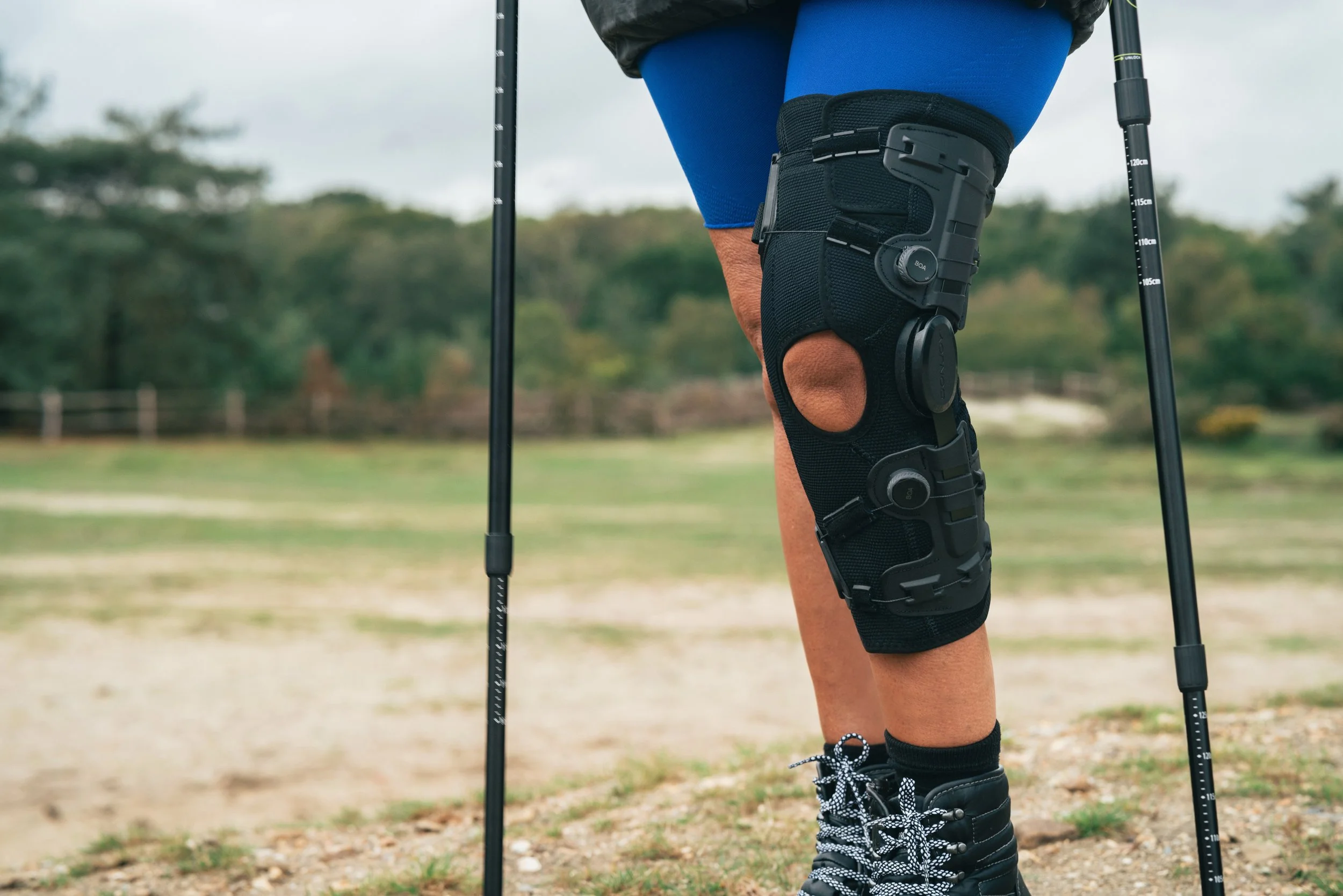 A person wearing a knee brace with an open hole, black walking shoes with white shoelaces, blue water-resistant shorts, and holding trekking poles, standing outdoors on a dirt path with trees in the background.