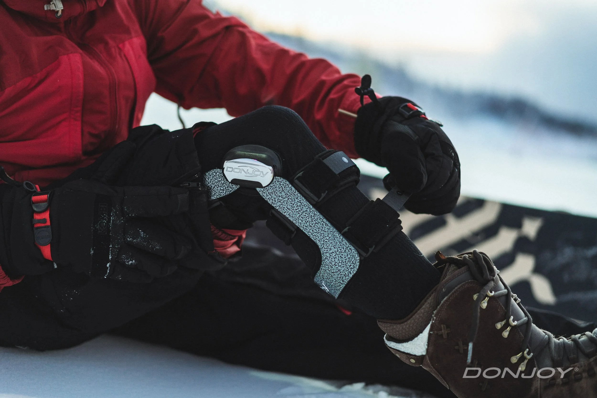 A person wearing a red jacket and black gloves is adjusting his knee brace while sitting on snow in a winter landscape.