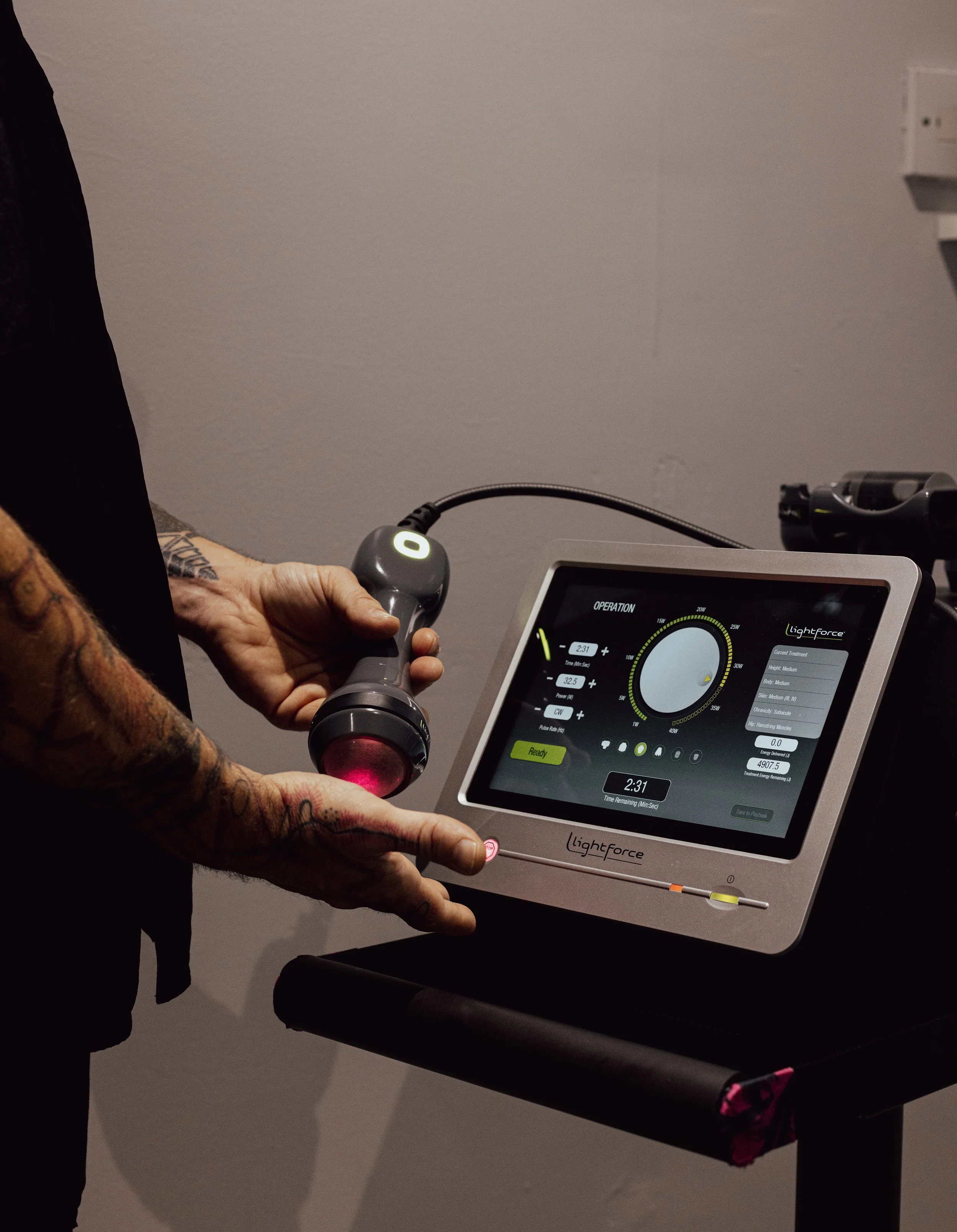 Person holding the high intensity laser therapy device connected to the main machine with a touchscreen control panel displaying settings, on a black table.