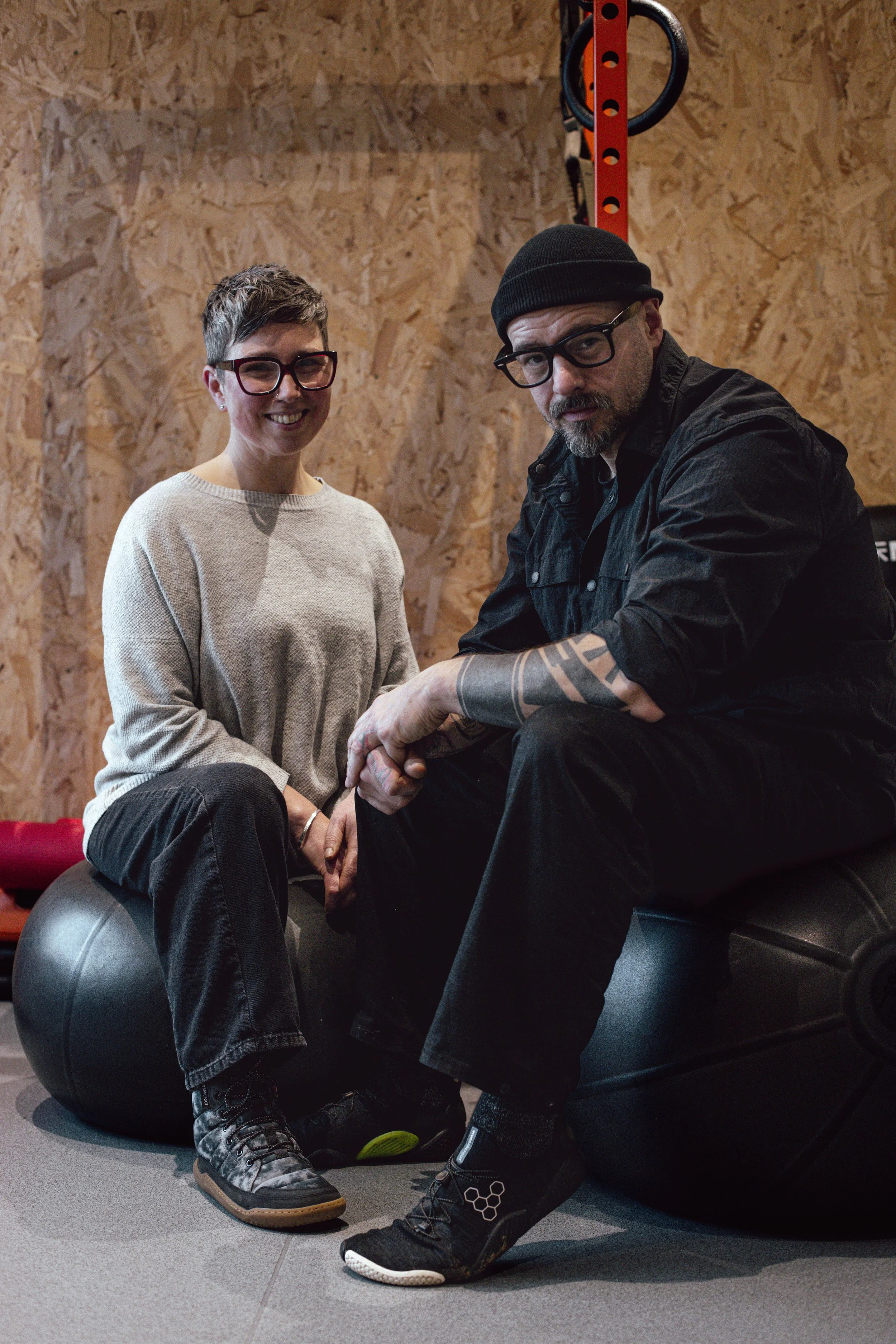 A woman with short hair, glasses, and a beige sweater sitting on a therapy ball, smiling. A man with a beard, glasses, a black beanie, black shirt, and tattoos on his arm, sitting on a therapy ball, in a gym or therapy space with plywood walls and fitness equipment.