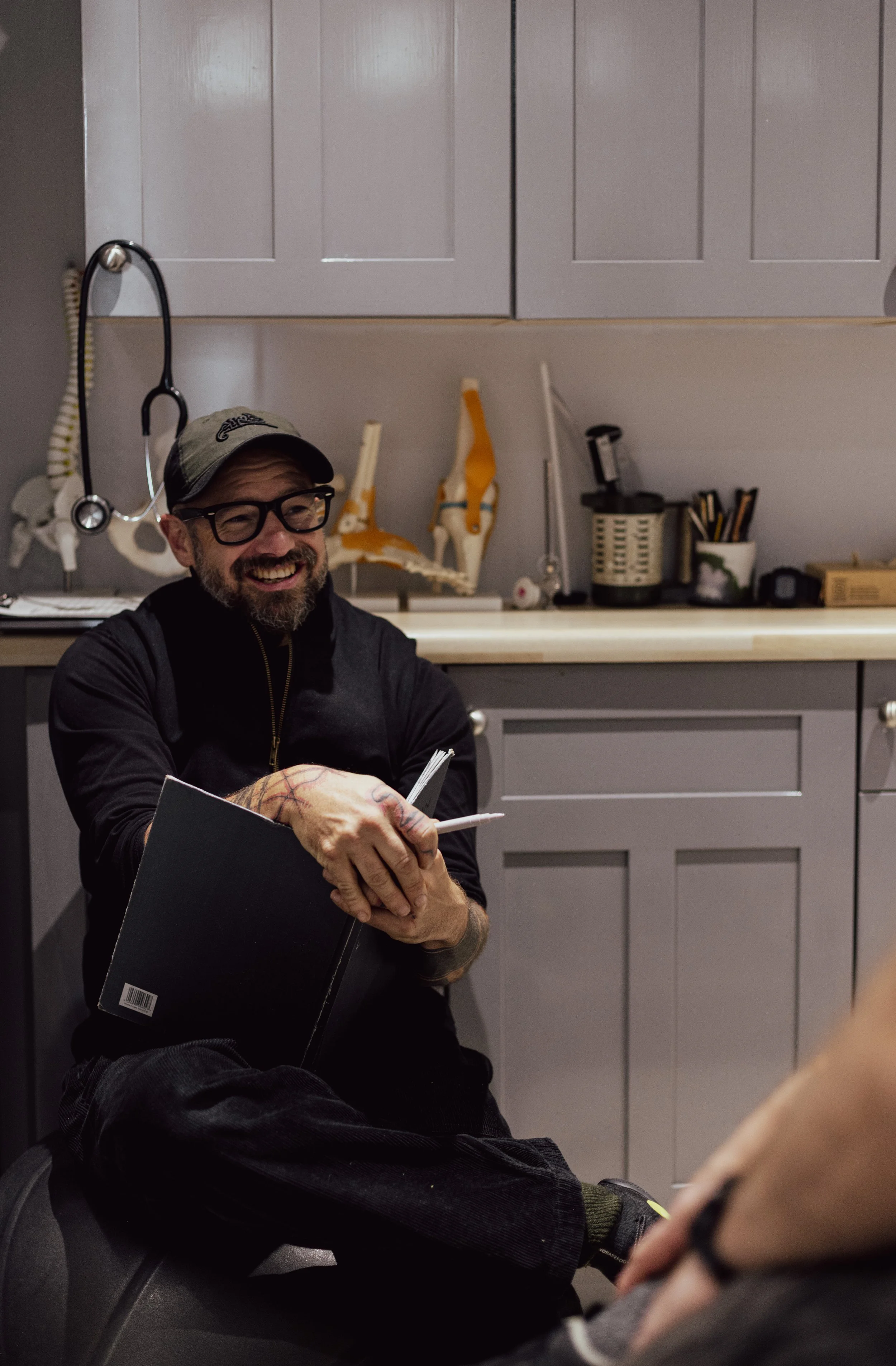 A man with glasses and a beard, smiling, sitting in a room with medical supplies, holding a black click board for a initial assessment, and wearing a cap and black clothing.