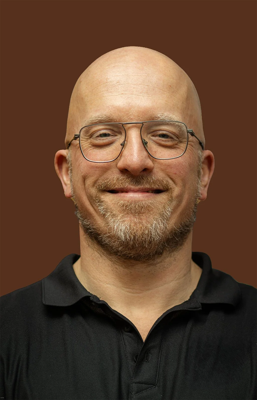 A smiling man with a bald head, glasses, and a goatee, wearing a black collared shirt, standing in front of a plain brown background.