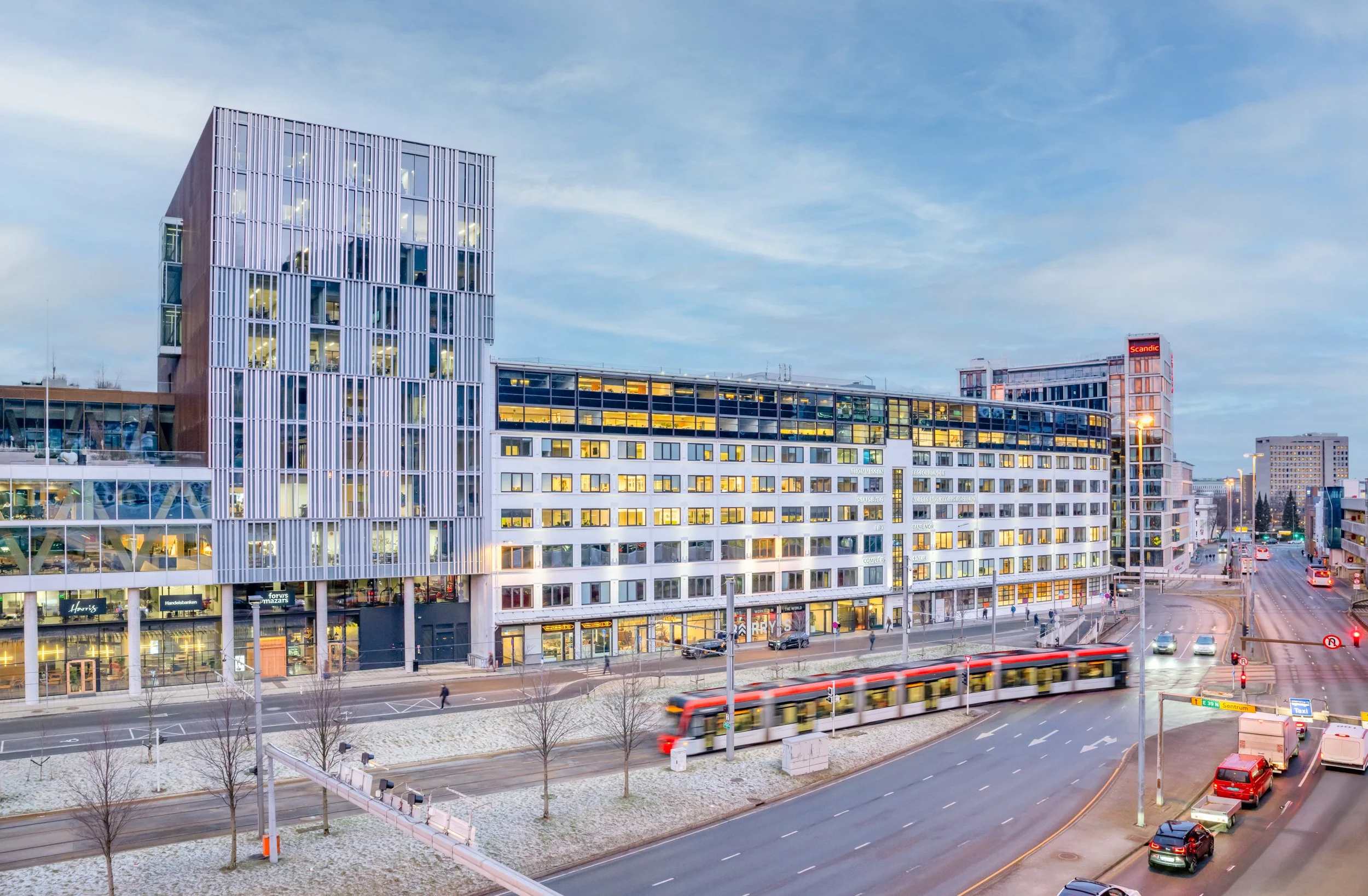 Bygate med moderne bygninger, trikk på veien, biler og folk som går, tidlig kveld med opplyste vinduer, blå himmel med spredte skyer.