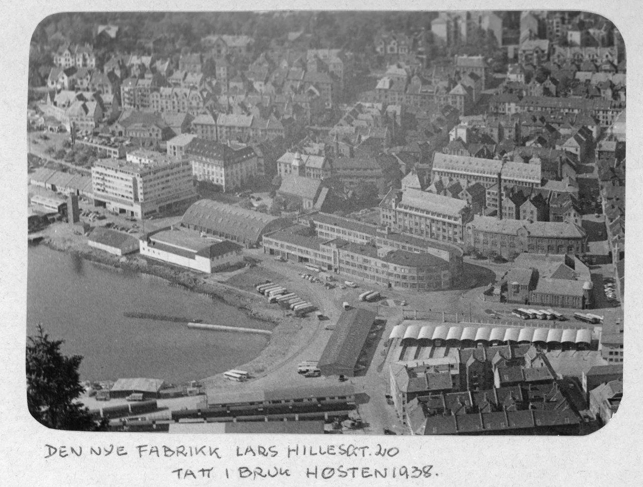 En svart-hvitt foto av en by med flere bygninger, inklusive en fabrikk nær vannet i forgrunnen, med en rekke biler parkert og en jernbanestasjon med tog. Notat under bildet indikerer at det viser den nye fabrikken Lars Hillesgat, tatt i bruk høsten 1938.