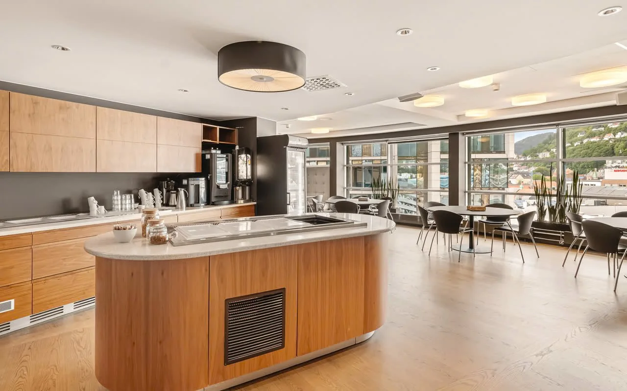 Et moderne kaffebarområde med en rund kjøkkenøy i tre, store vinduer som slipper inn naturlig lys, og flere spisestoler rundt vinduene.