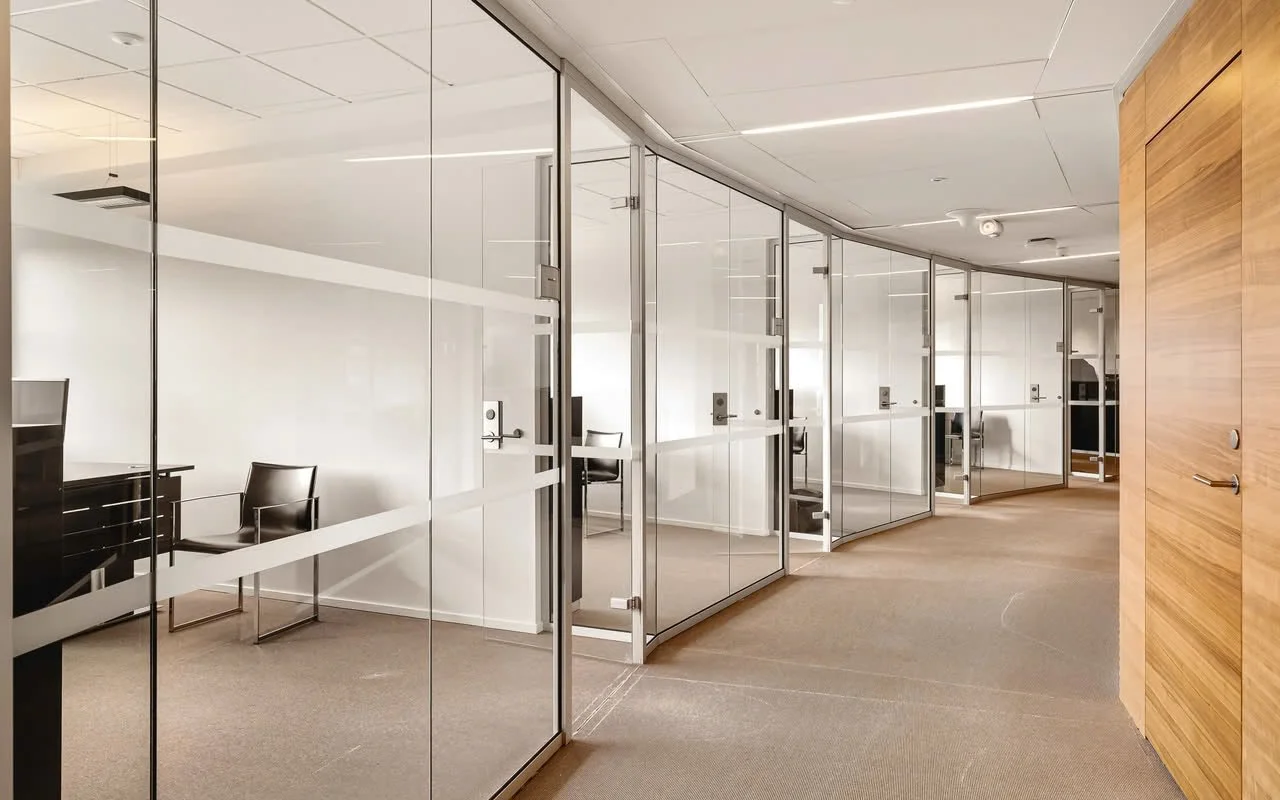 Kontor med private arbeidsstasjoner, adskilt av glassvegger, i et moderne, lyst rom med tre-paneled vegg og beige teppe.