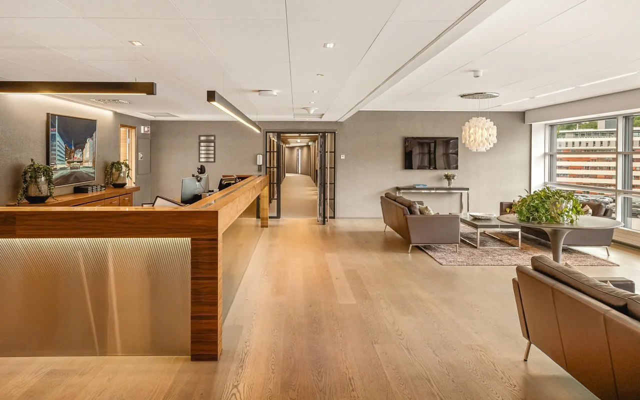 Lenge velkomstområde i en moderne bygning med sittegruppe, stor vindu, og dekorative planter.