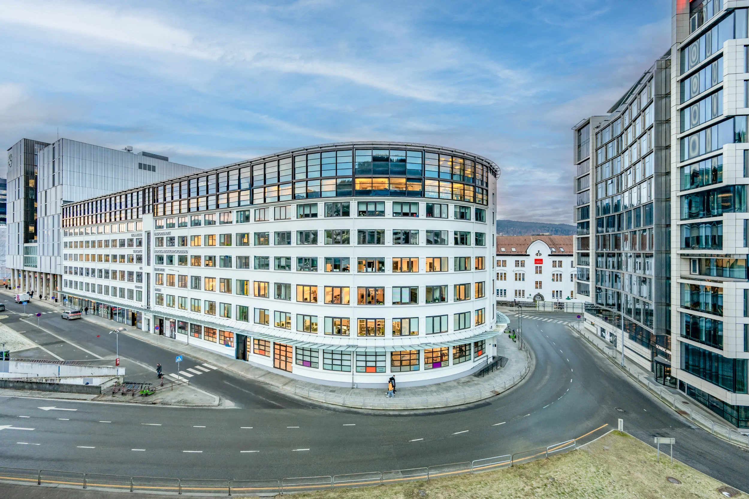 Moderne kontorbygninger langs en svingete vei, noen personer går på fortauet, med en blå himmel og skyer i bakgrunnen.