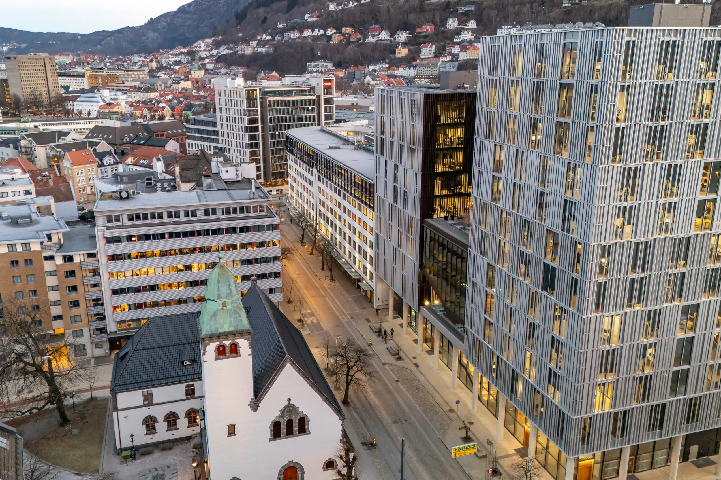 Bybilde med moderne kontorbygninger og en eldre kirke i forgrunnen, med hus og fjell i bakgrunnen.