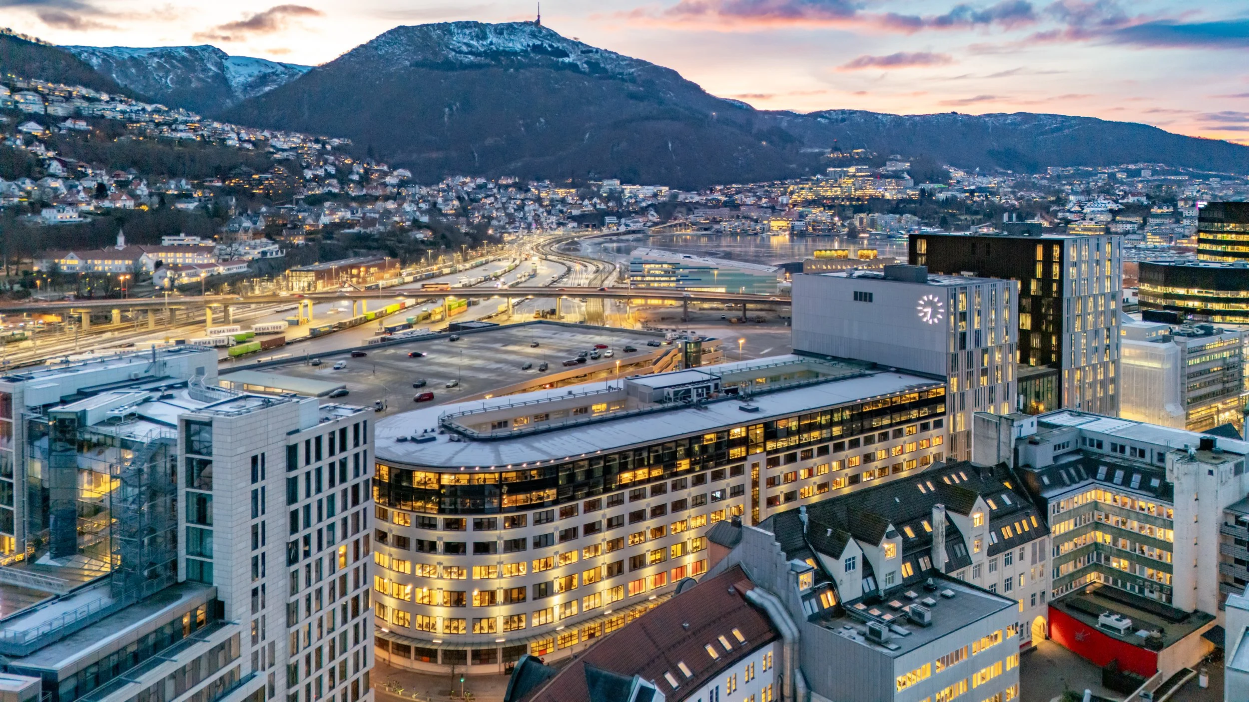 Utsikt over Bergen by med høye kontorbygninger, togstasjonen med tog og belysning, og fjell i bakgrunnen ved solnedgang