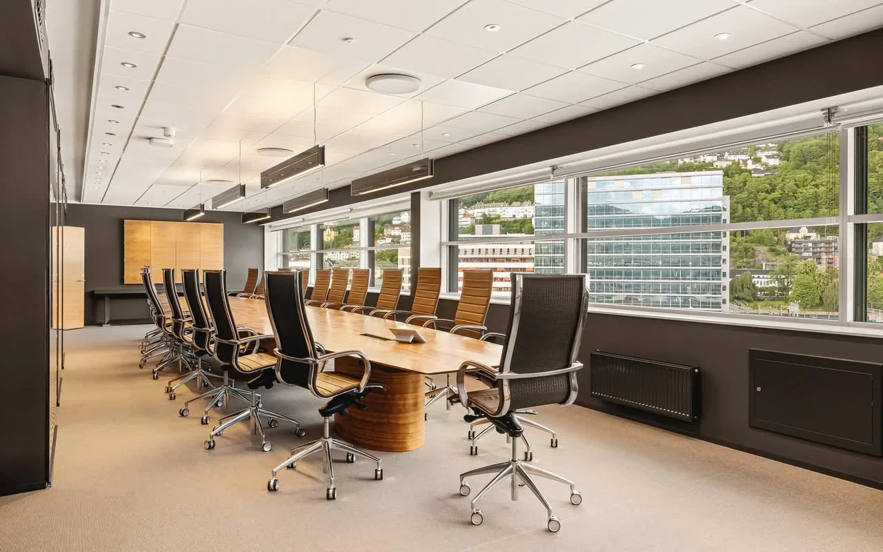 Et møterom med lang konferansebord og mange kontorstoler, store vinduer med utsikt over byen og grøntområder, og moderne belysning.