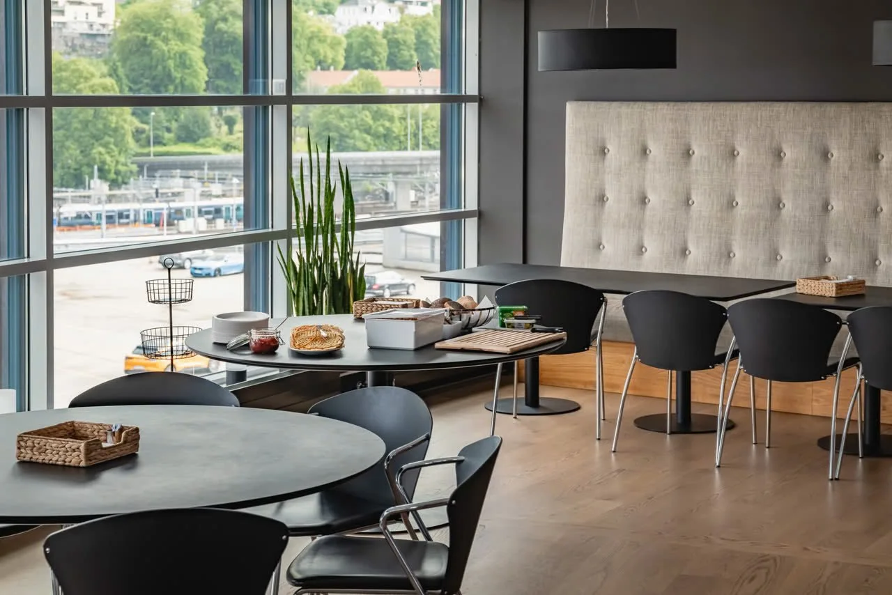 Innendørs spisestue med store vinduer, svarte bord og stoler, et oppdekket bord med mat og servise, en plante, og en sofa med hodestøtte på veggen.