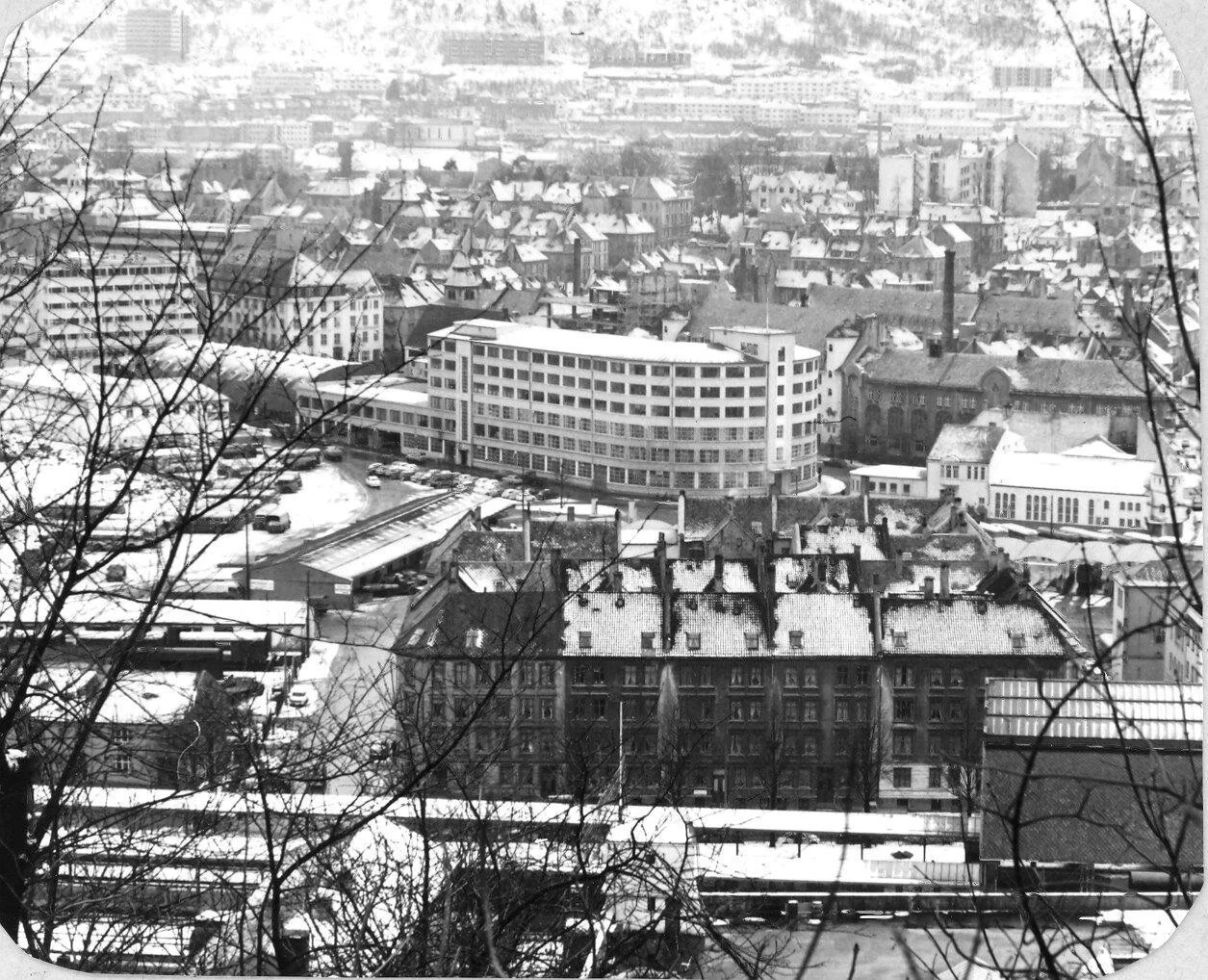Utsikt over en by i vinter med snø på takene og bakken, gamle og moderne bygninger, trær uten blader i forgrunnen.