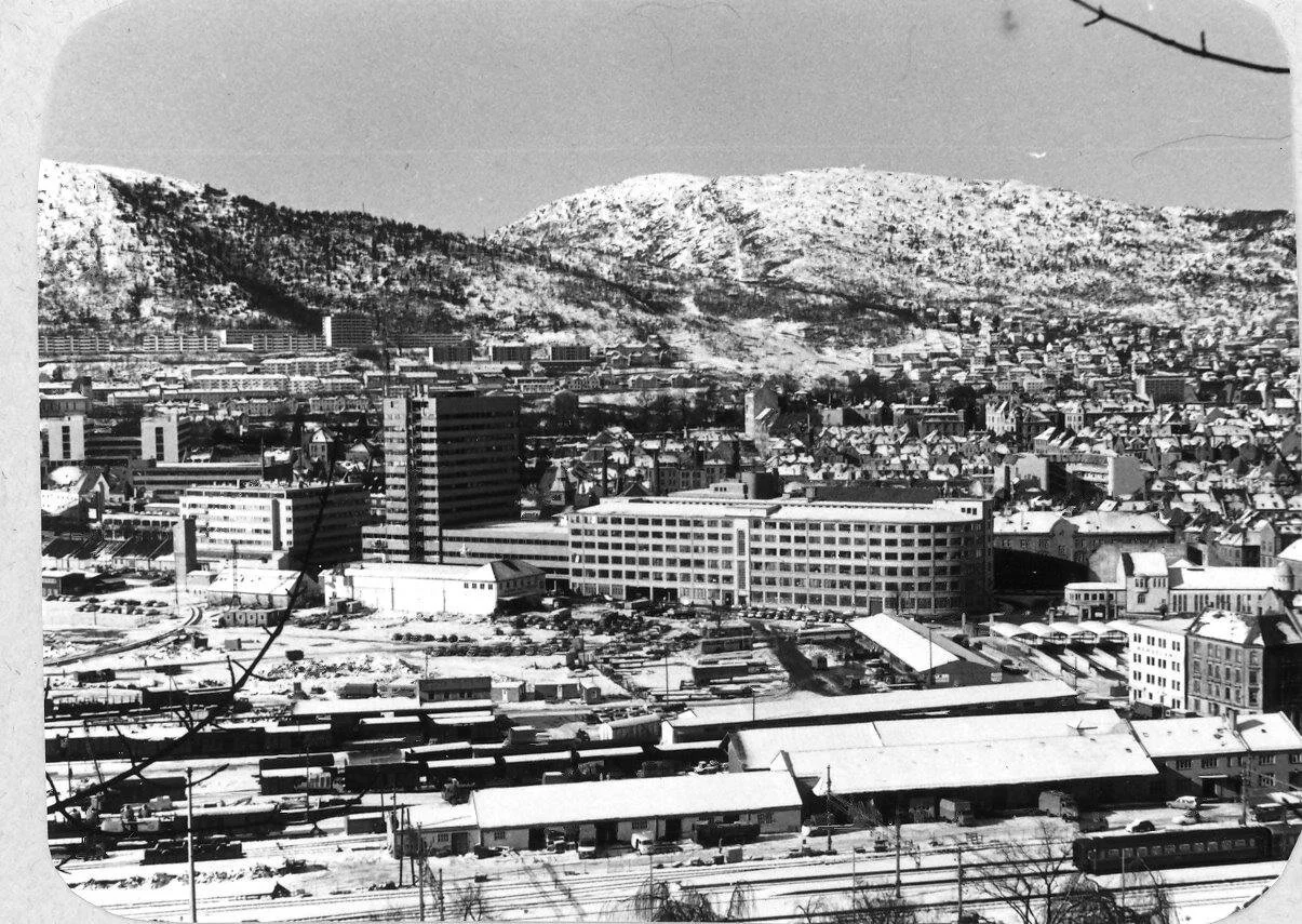 Sort-hvitt bilde av en by med fjell i bakgrunnen, inkludert flere bygninger, et togspor, og snødekte hustak.