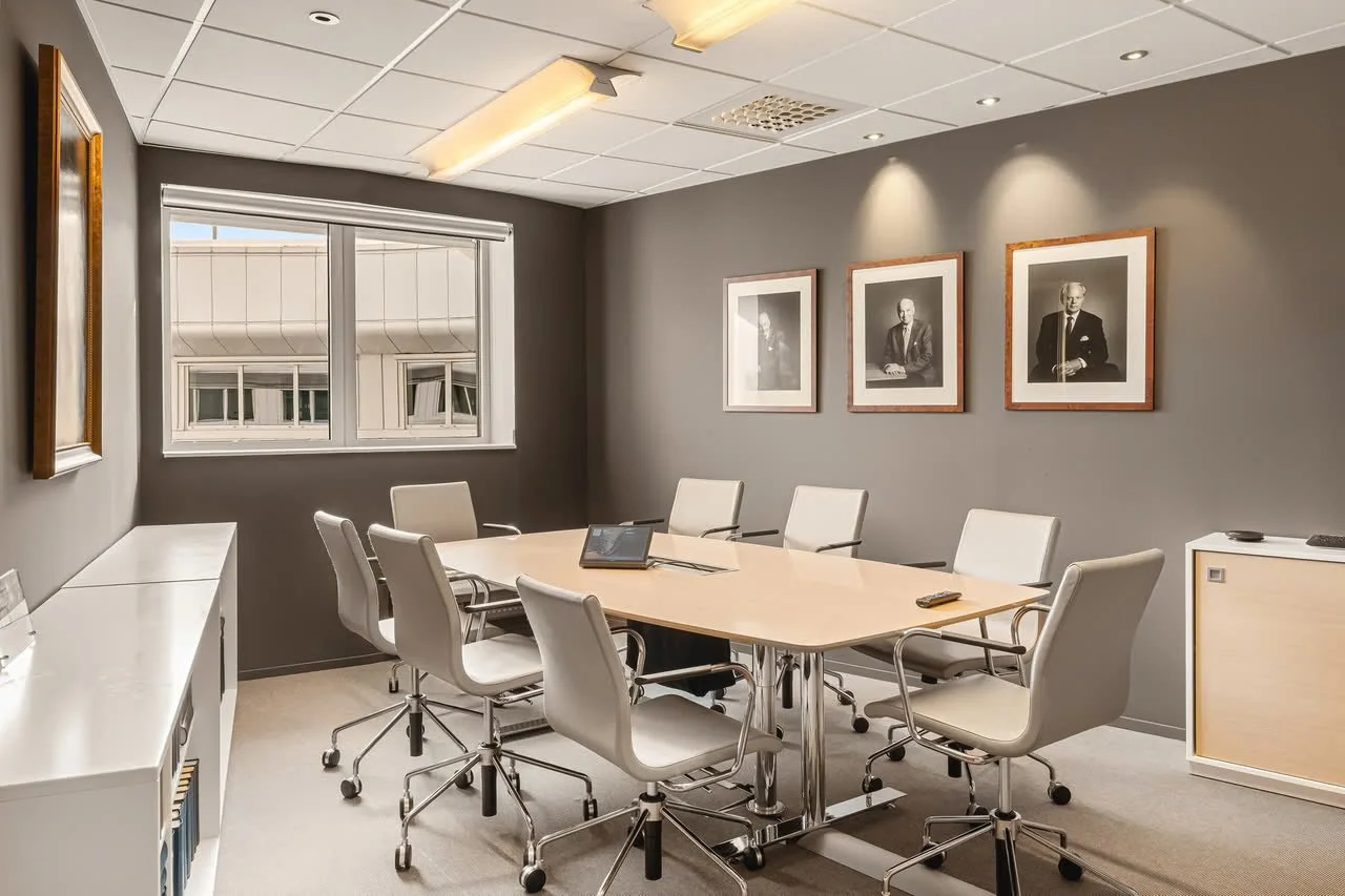 Møterom med rundbord, åtte hvite kontorstoler, veggdekor med tre portretter, vindu, taklys, og skap.