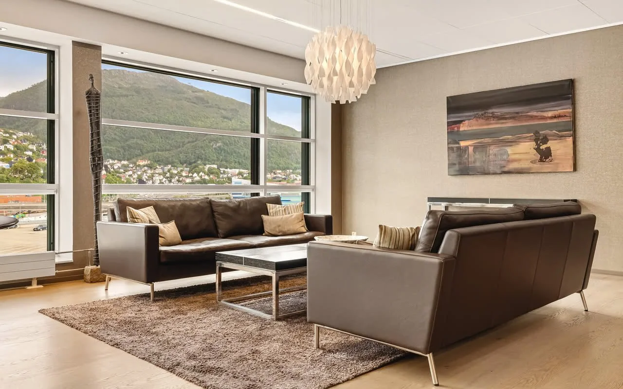 Stue med store vinduer, to svarte sofaer, et teppe, et lavt bord, veggkunst og en taklampe.