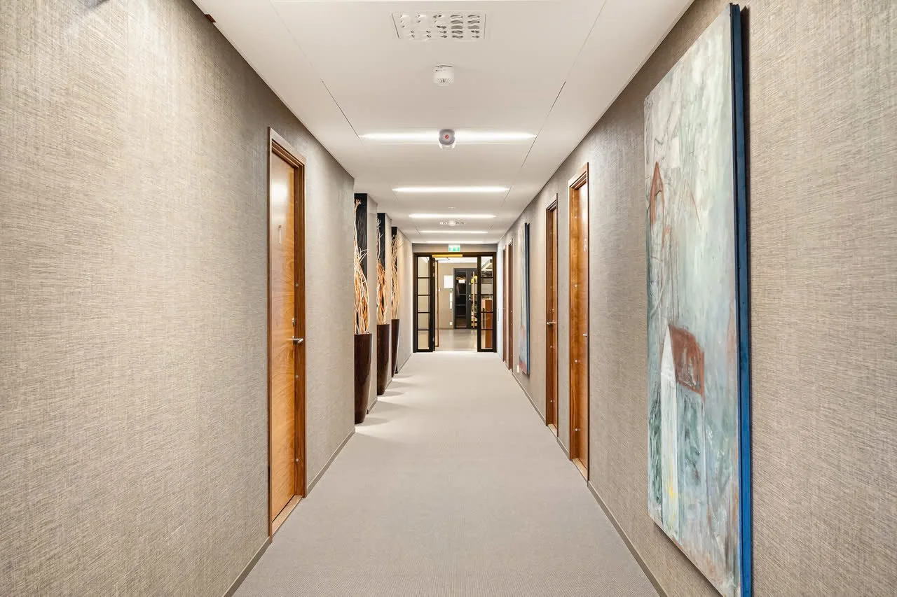 En lang, godt opplyst gang i en bygning, med flere dører på begge sider og abstrakte bilder på veggene, samt store blomsterpotter med dekorative grener.