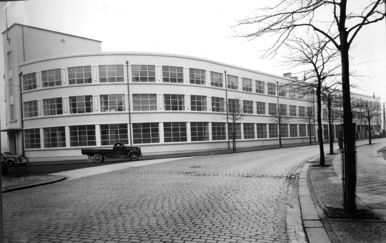 En svart-hvitt bilde av en bygning med mange vinduer og en rund gate foran, uten folk, med noen tomme parkerte biler og trær uten blader.