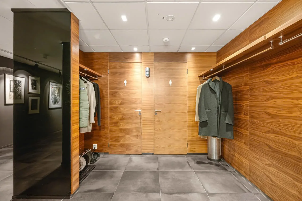 Et rom med tre parkasere i garderobeskap på veggen, tre par sko på gulvet, en vannkran på gulvet, og et par mænd til venstre i bildet, som henger på en vegg med bilder.