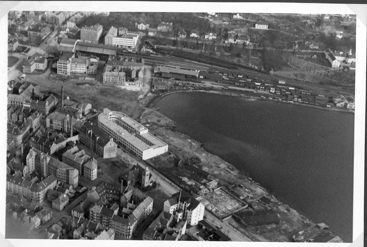 Sort/hvitt luftfoto av en by med vannkropp, jernbanespor og mange bygninger, fra en høyde