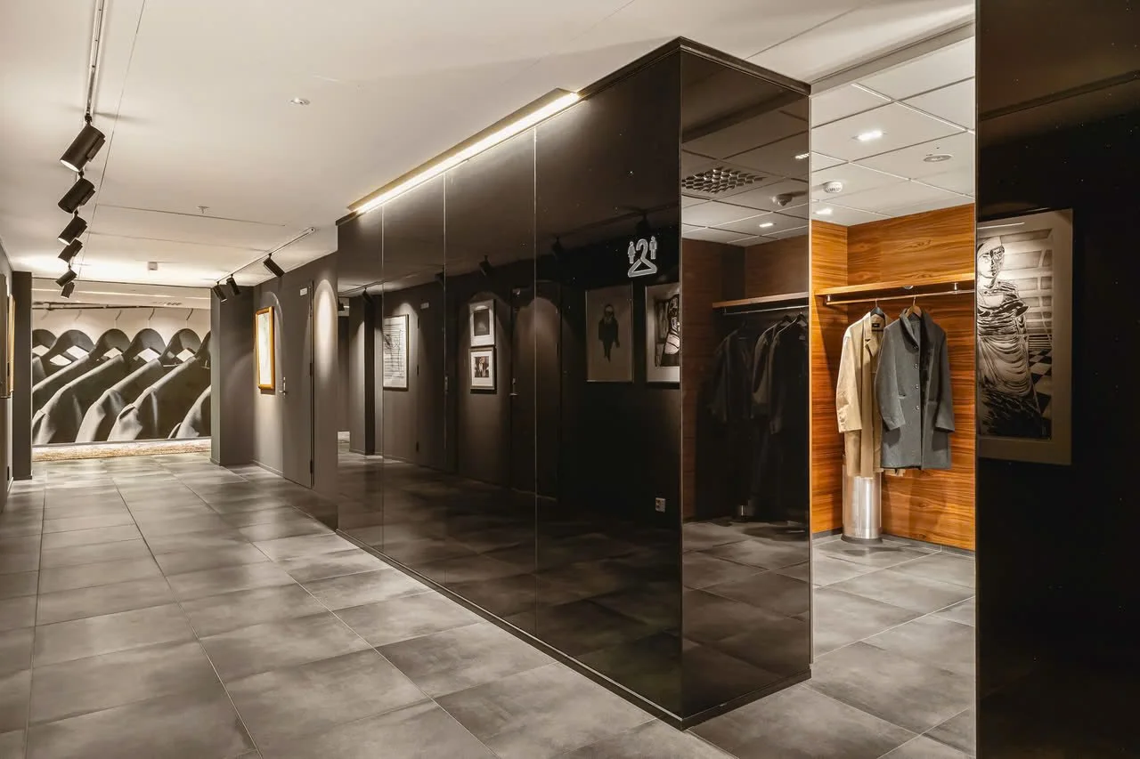 Interiør av en butikk eller et galleri med klær og kunst, mørke vegger, svart og trepaneler, fotografier på veggene, og en krok med klær.