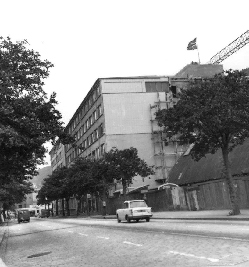 En svart-hvitt gate med biler og et fleretasjesbygg, trær langs fortauet, og en byggekran med et flagg i bakgrunnen.