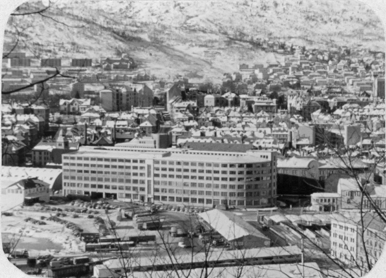 Sort-hvitt bilde av en by med mange bygninger, med en stor, langstrakt bygning i forgrunnen og fjell i bakgrunnen.