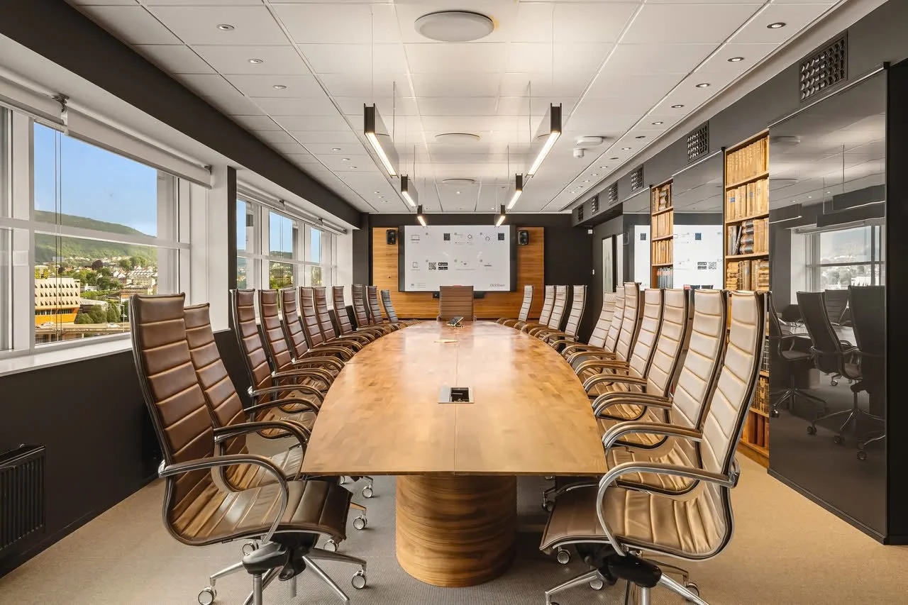 Møterom med lang trebord og mange komfortable kontorstoler, store vinduer med utsikt over byen og fjell, lysende lamper i taket, bokhyller med bøker