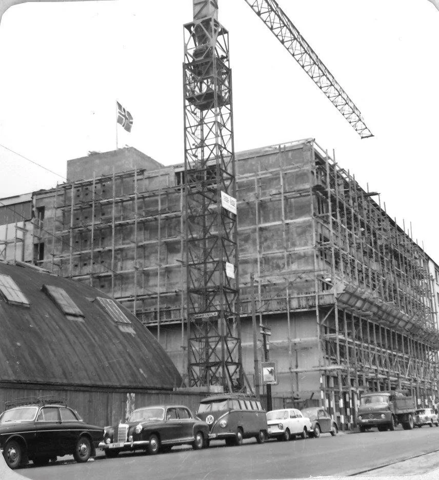 Byggearbeid med et flattløpt bygg under konstruksjon, en stor kran og flere biler parkert på gaten.