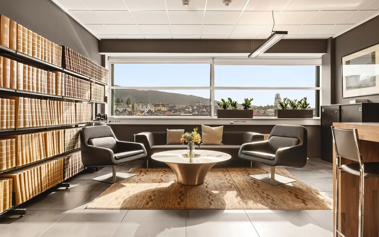 Lyse rom med store vinduer, to svarte lenestoler, en rund hvit kollapsbord med en blomsterpotte, en svart sofa med to puter, et skogkledt landskap i utsikten, og et parkettgulv med teppe.