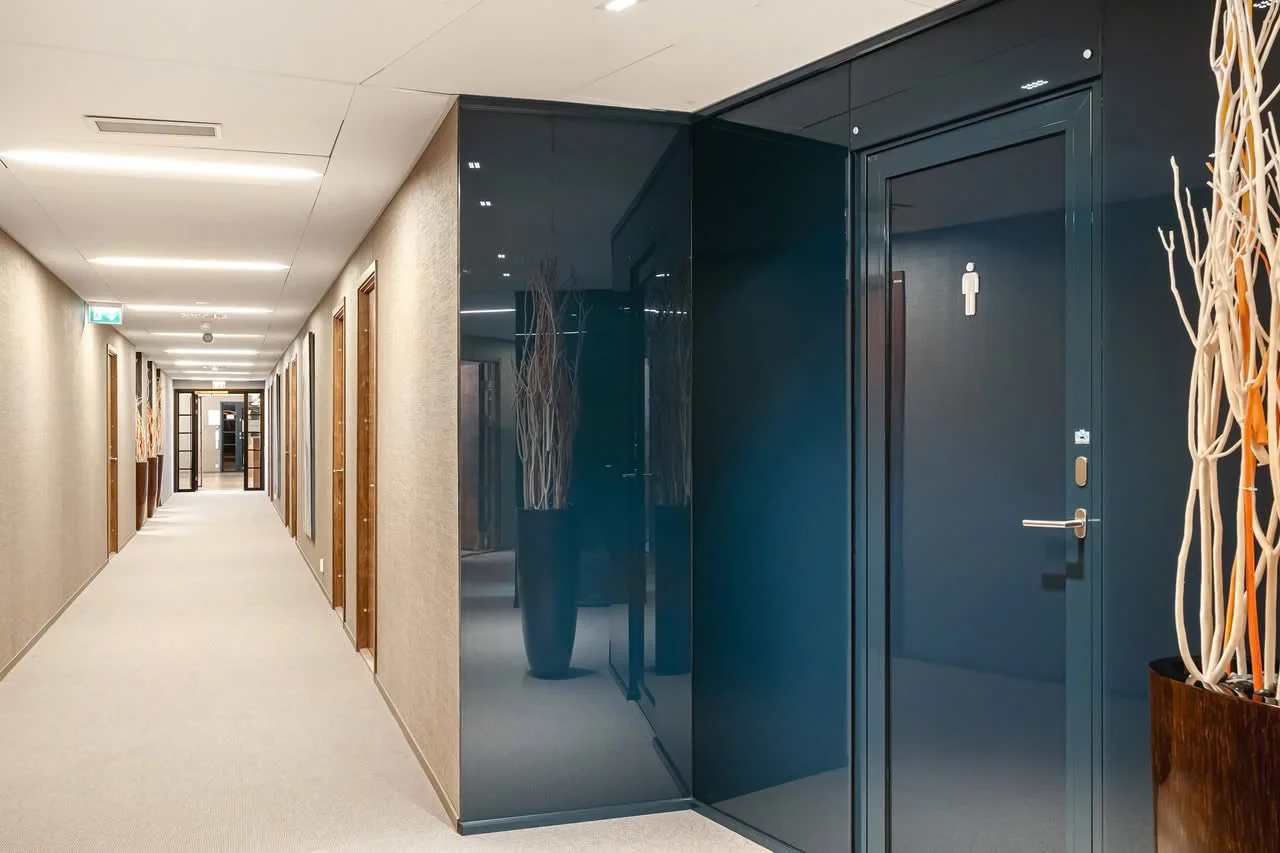 En lang hotell korridor med dører på begge sider og en ensfarget vegg med noen dekorative elementer. På høyre side er en mørk blå dør med et toalett-symbol.