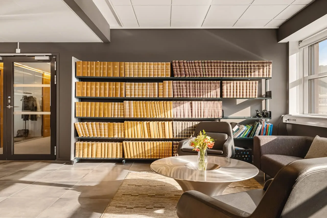 Et kontor med en svart vegg, to hyller med bøker i beige og falmet brunt, et bord med en vase med blomster, to stoler og en sofa. Sollys lyser inn gjennom et vindu på høyre side.