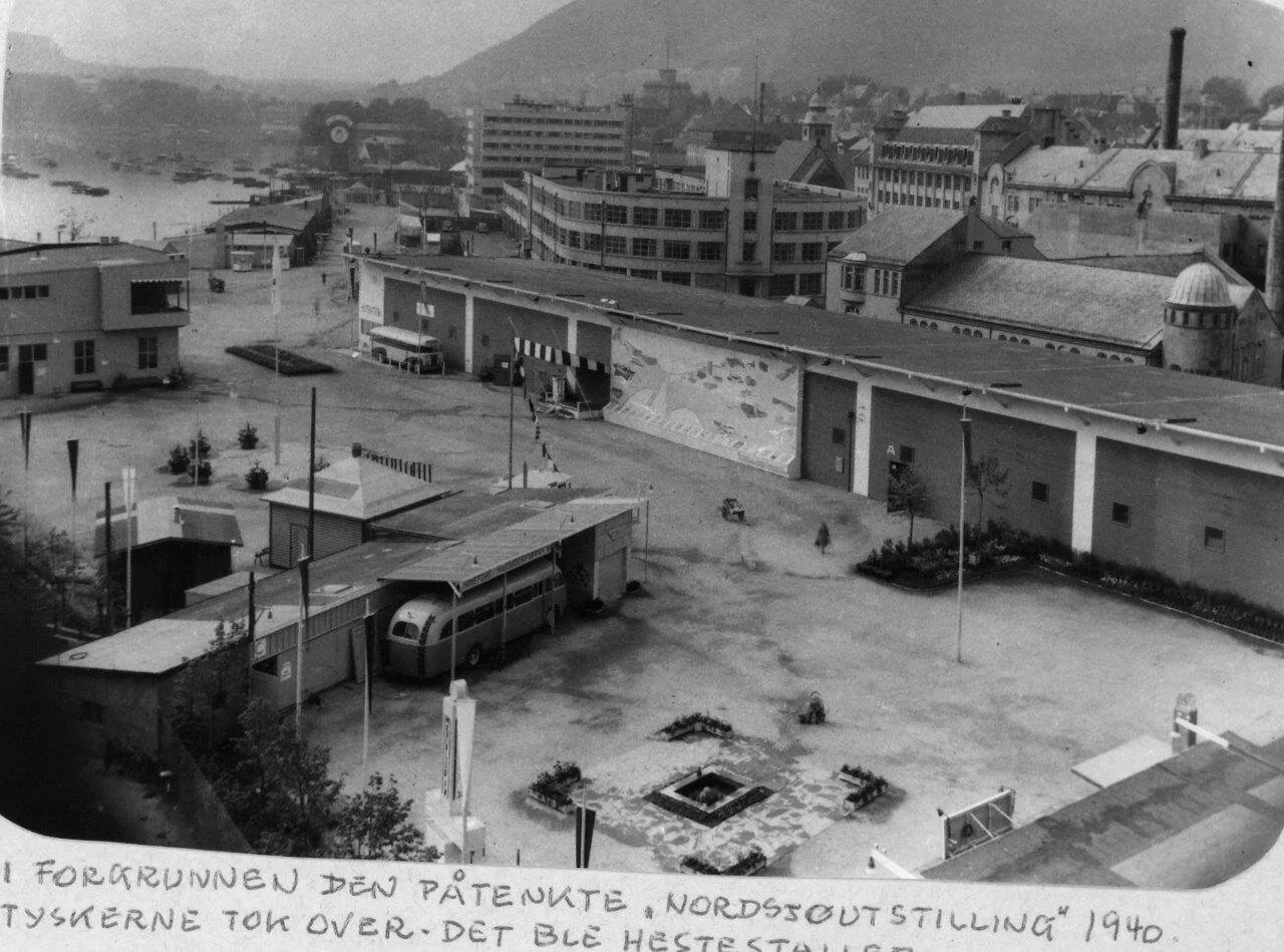 Sort-hvitt bilde av et bylandskap fra 1940-tallet, med mange bygninger, en fergeterminal og et busstopp, samt en liten grønn plass med planter og en vannfontene.