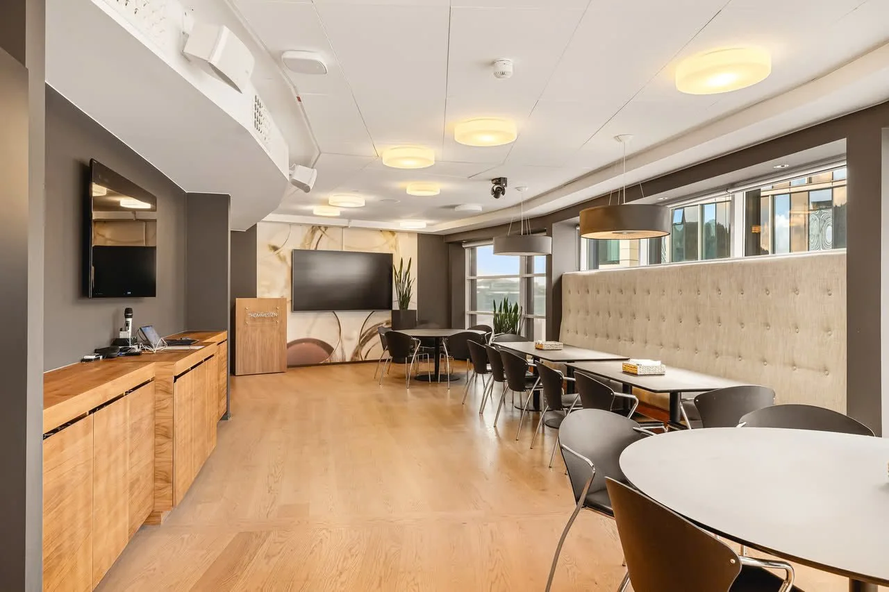 Interiør av et moderne møte- eller konferanserom med stoler og bord, TV-skjerm på veggen, store vinduer og moderne belysning.