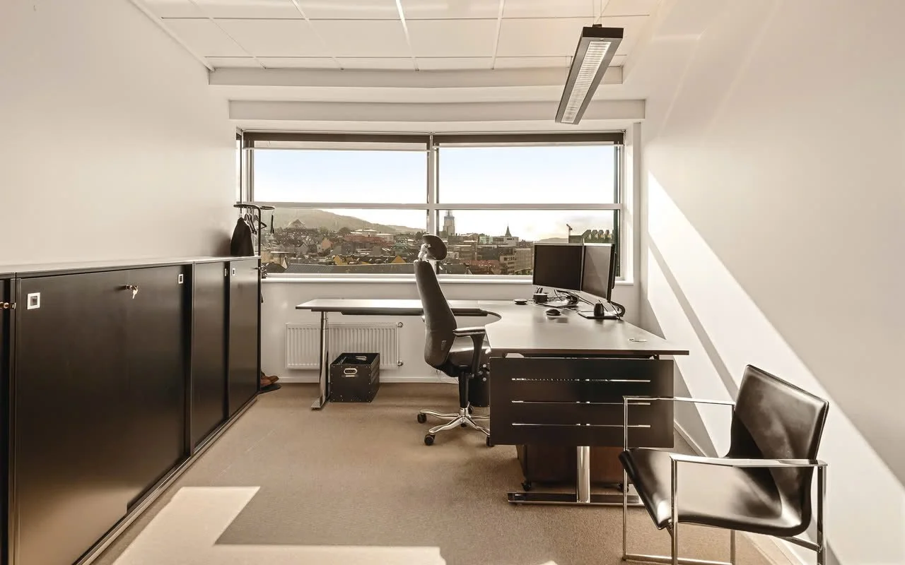 Lite kontor med skrivebord, kontorstol, og dataskjermer foran et vindu som viser en by og fjell i bakgrunnen.