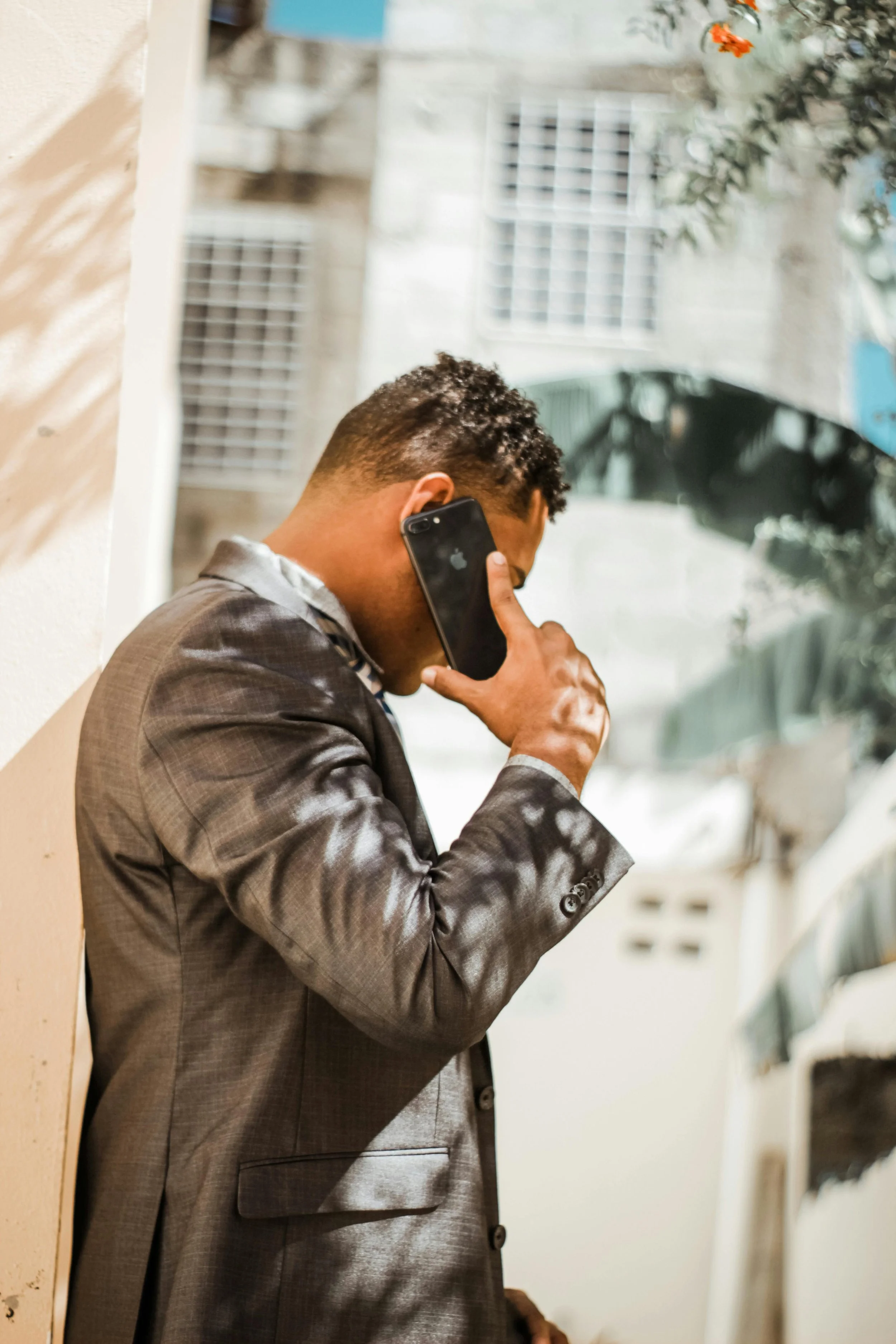 A man in a gray checkered suit is talking on a black smartphone, standing against a wall with a blurred building in the background.